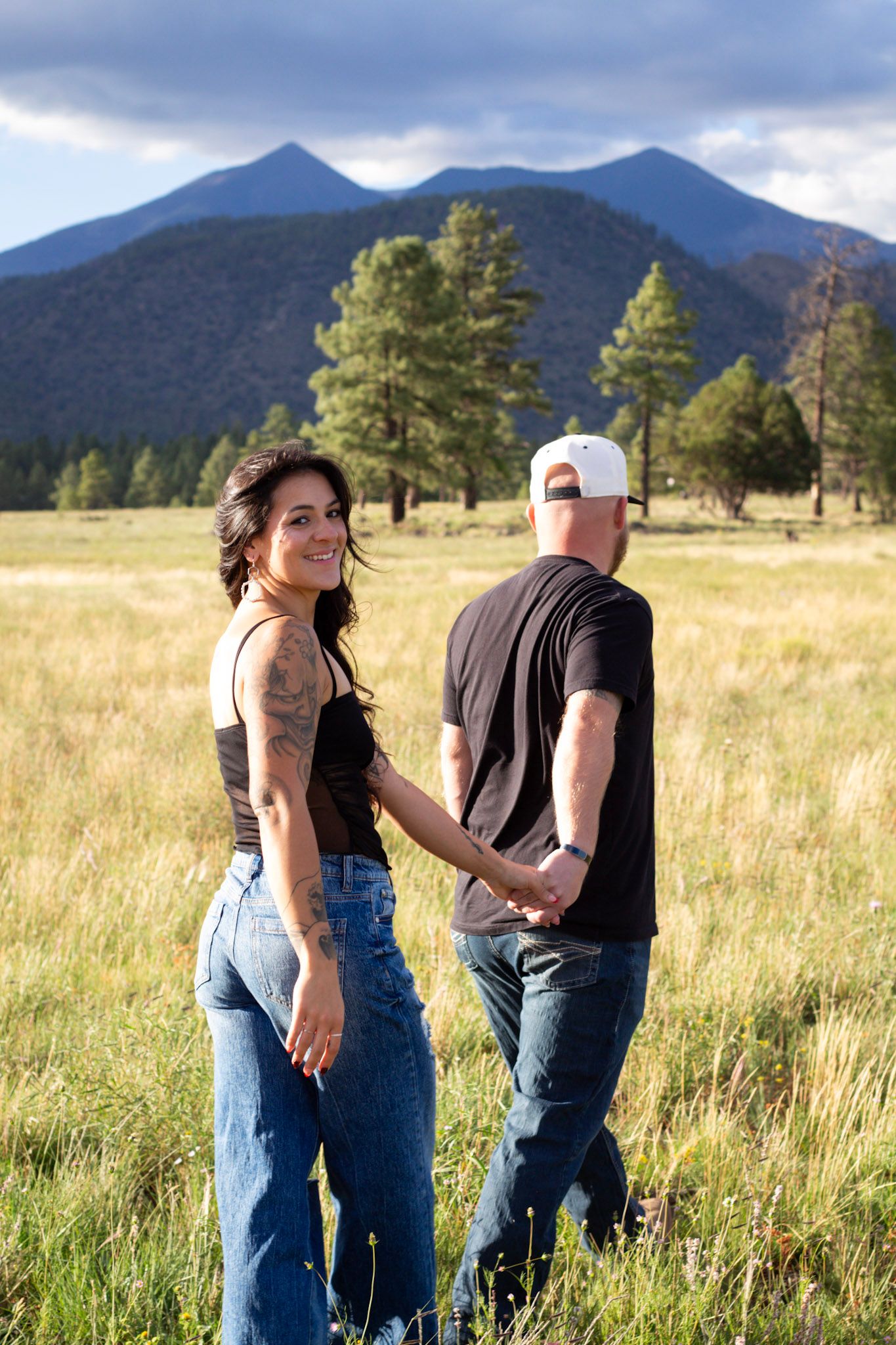 Couple holding hands in a sunny mountain meadow, woman smiling back at camera, pine trees and peaks behind.