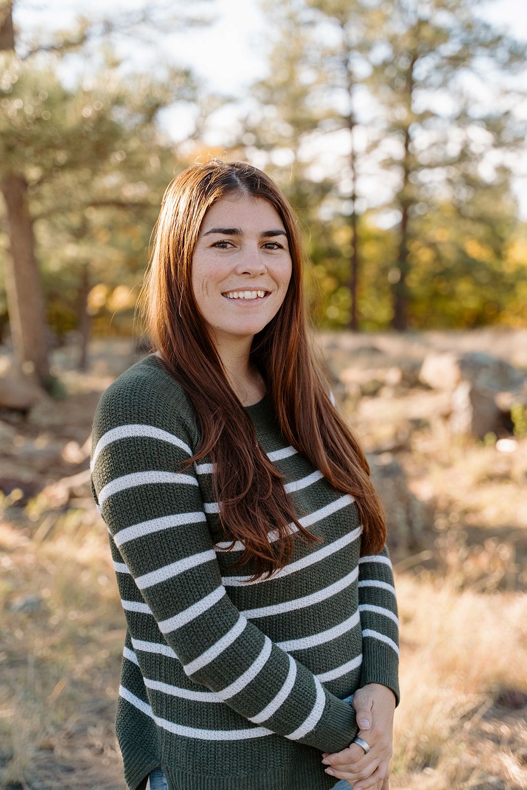 Young woman with long auburn hair smiling outdoors in a green striped sweater among autumn trees.