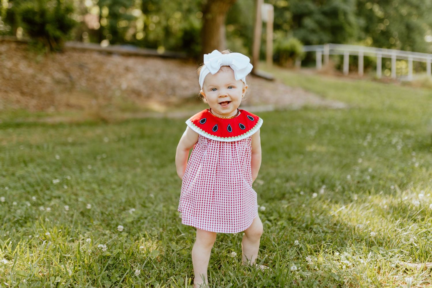 Smiling toddler girl in red gingham watermelon dress with white bow standing on green grass outdoors.