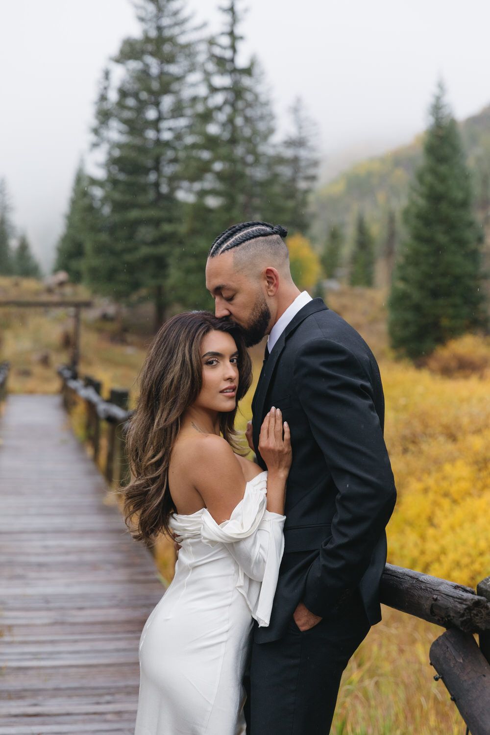A couple in wedding attire embraces on a wooden bridge surrounded by autumn foliage and evergreen trees in a misty mountain setting.