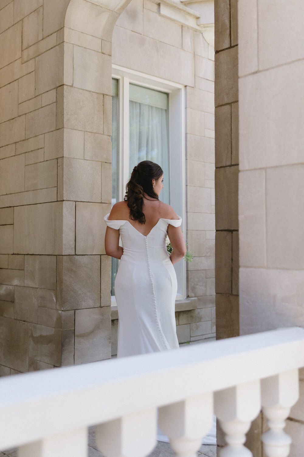 A bride in an elegant white off-shoulder wedding dress stands next to stone columns and white railings at her atlanta courthouse elopement