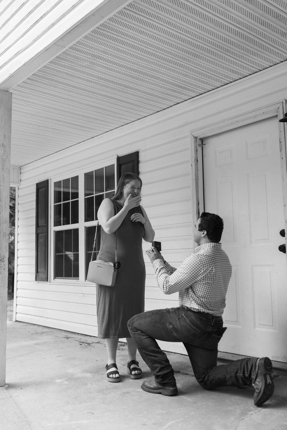 Man kneeling on one knee proposing to woman on porch in black and white photo.