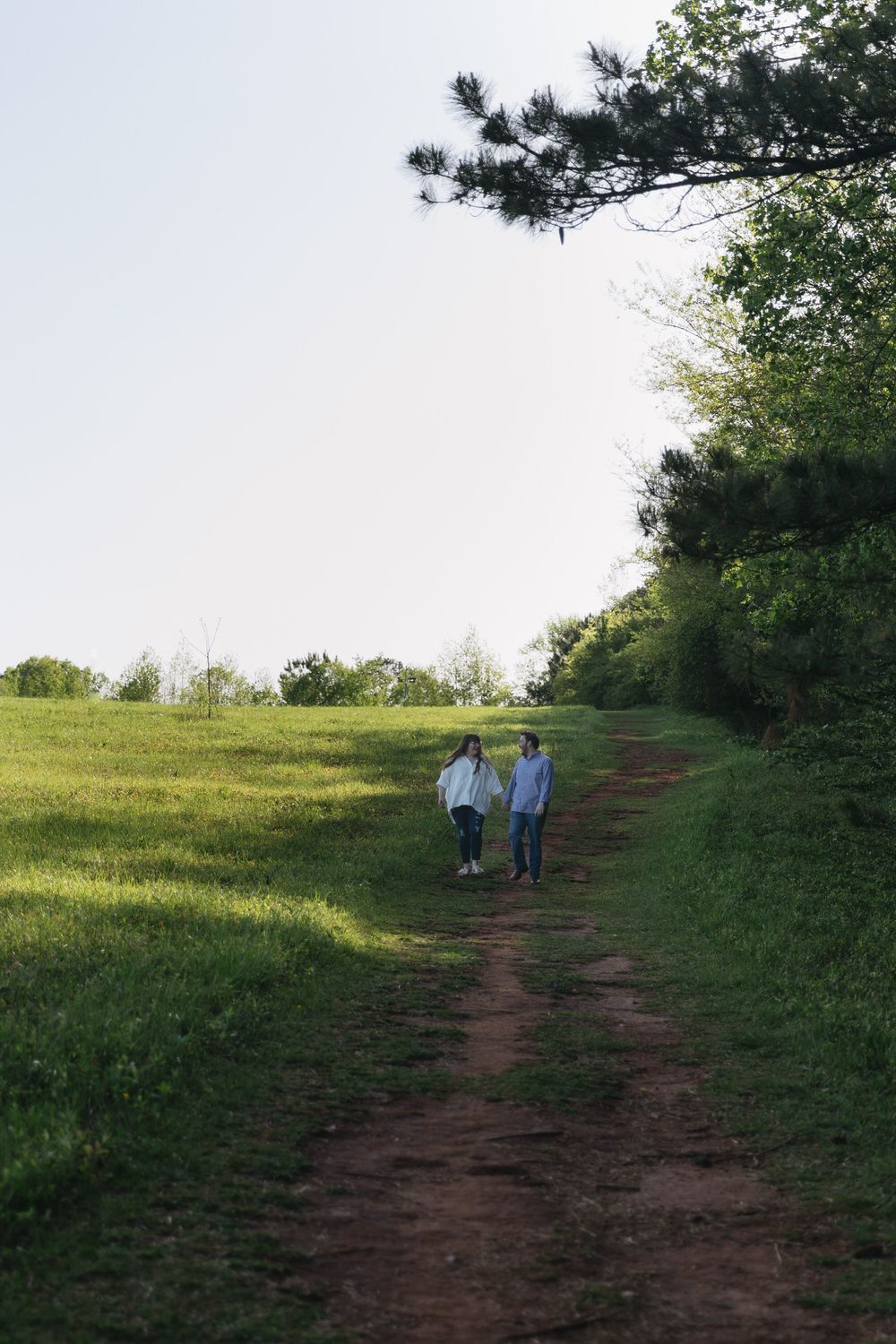 A couple walks along a winding dirt path through a grassy hillside bordered by pine trees on a serene afternoon in north georgia