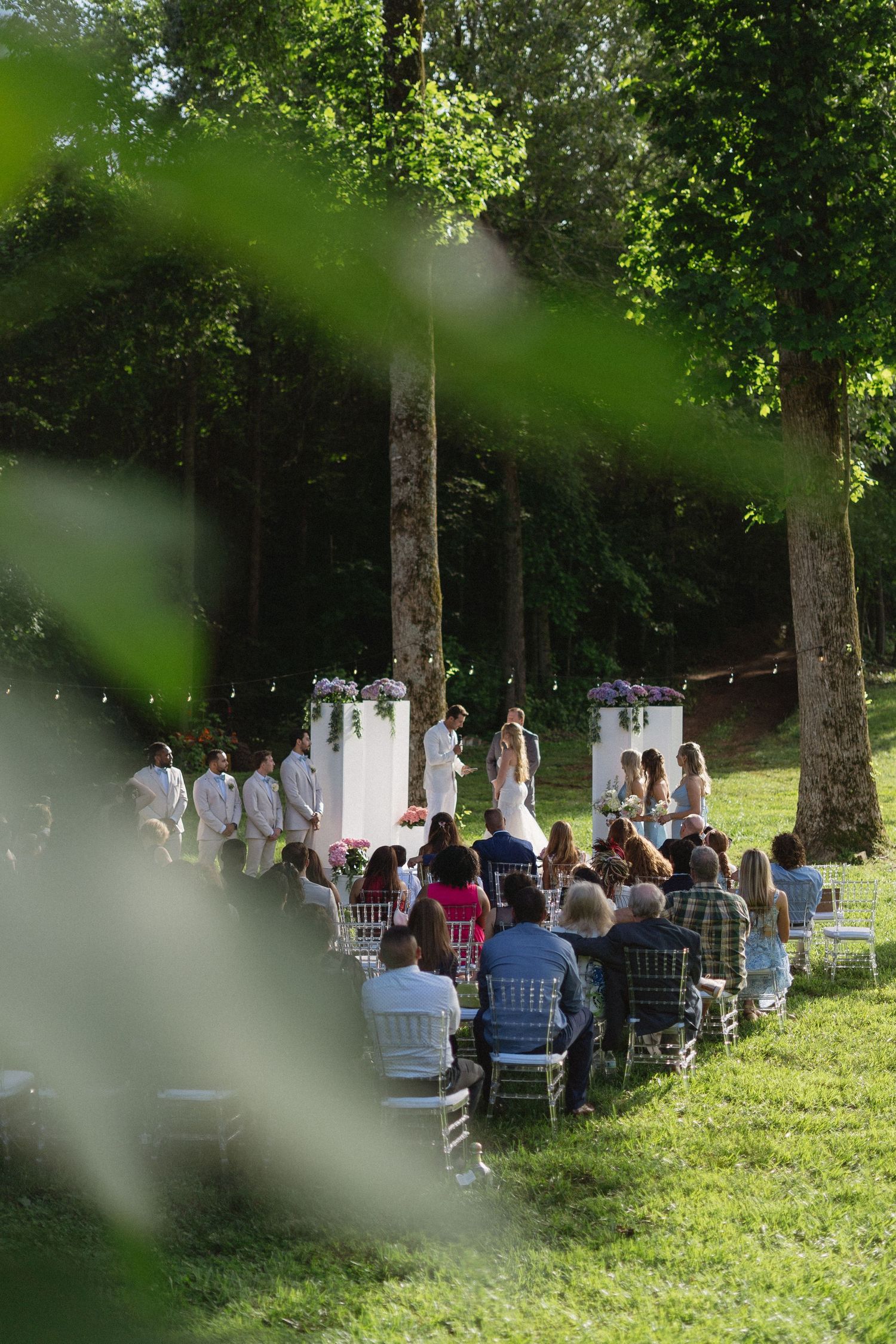 Outdoor garden wedding ceremony setup with white chairs arranged on a grassy lawn beneath tall trees in natural lighting.