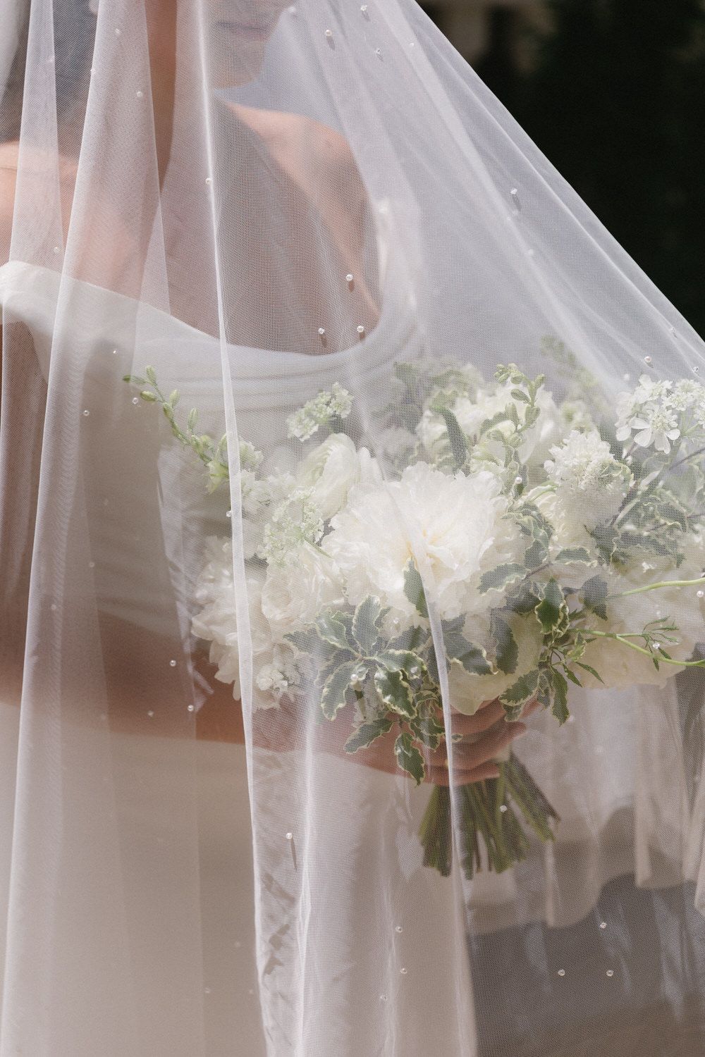 A white bridal bouquet with soft florals is held beneath a sheer wedding veil in an ethereal composition at bride's Denver elopement