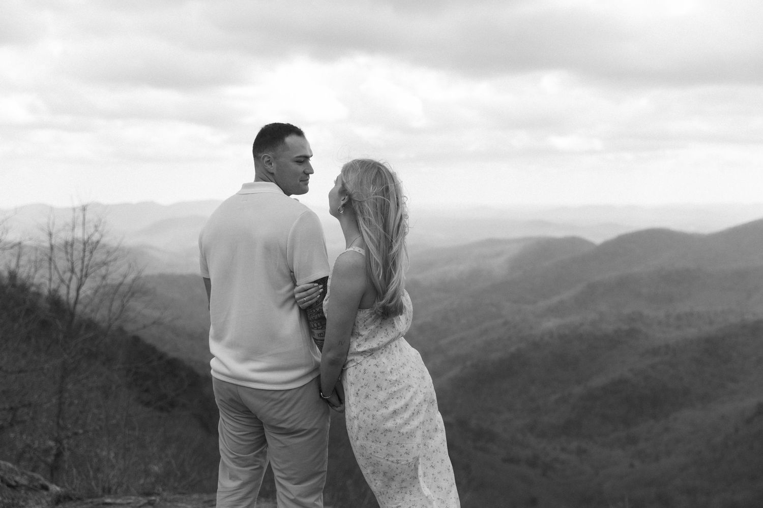 Black and white photo of a couple embracing on a mountain overlook with scenic rolling hills in the background.