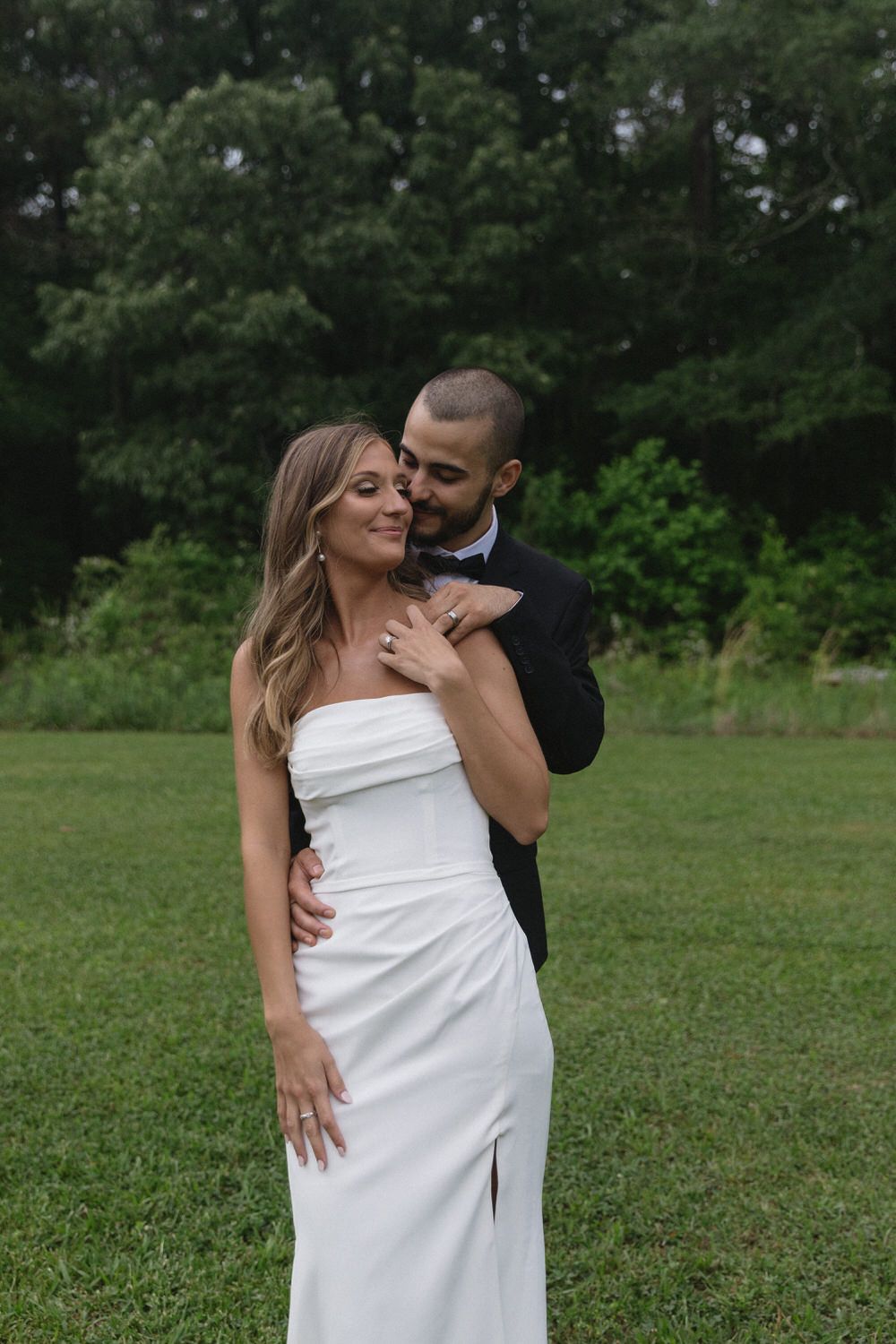 A happy couple embraces outdoors, with the bride wearing a strapless white gown and groom in black suit against green foliage.
