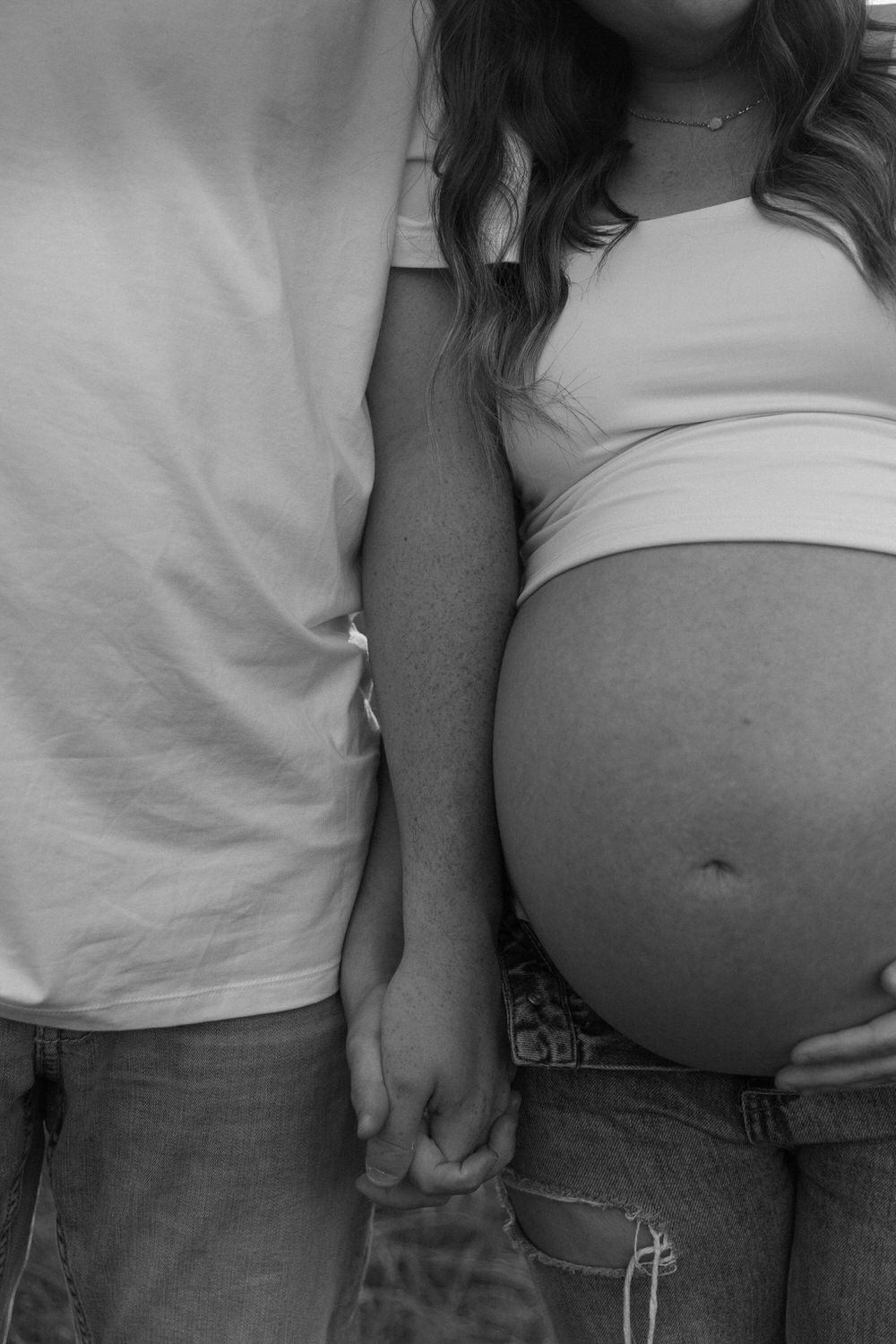 Black and white photo of a pregnant woman holding hands with her partner, showcasing her baby bump in jeans and a crop top.