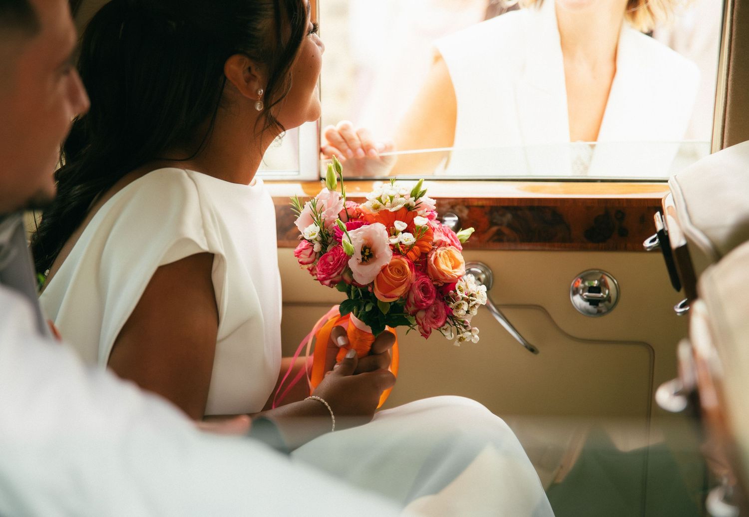 Une mariée élégamment vêtue tient son bouquet de fleurs aux teintes roses, orange et blanches tout en regardant par la fenêtre d'une voiture