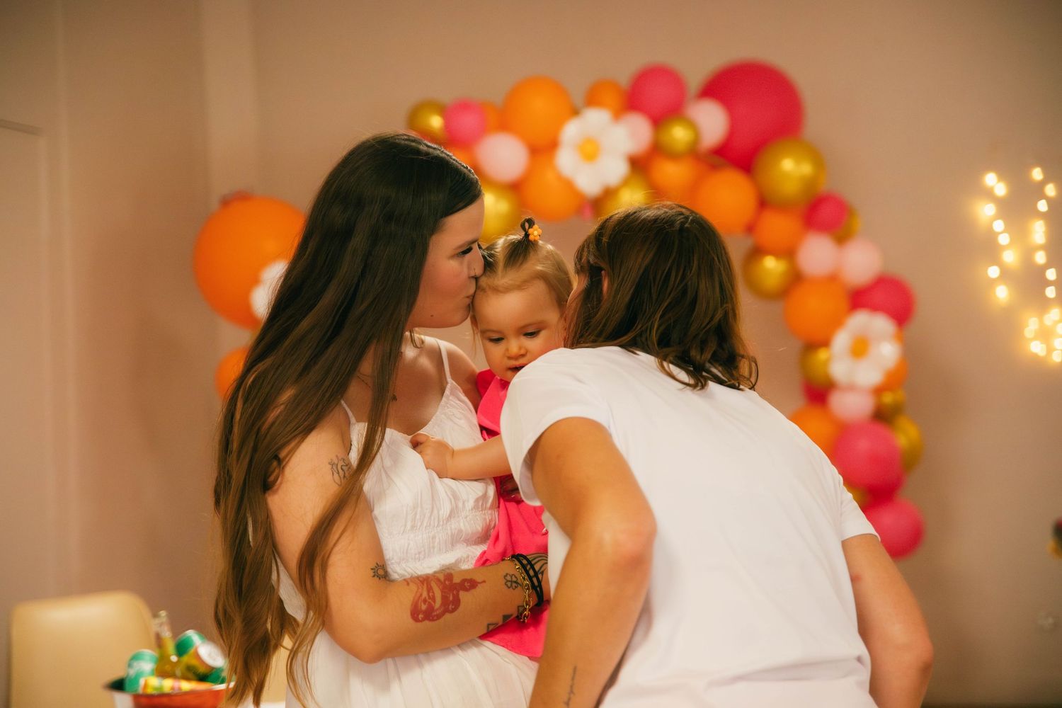 Maman et proches câlinant un bébé lors d'un anniversaire, arche de ballons orange et rose en fond. Reportage famille Essonne. Photographe 91