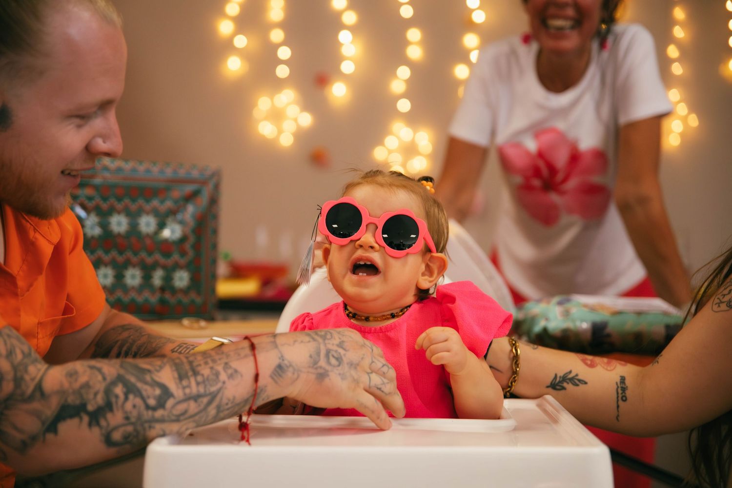Séance anniversaire Essonne 91, bébé hilare avec lunettes de soleil roses entourée de ses parents tatoués et guirlandes lumineuses, moment