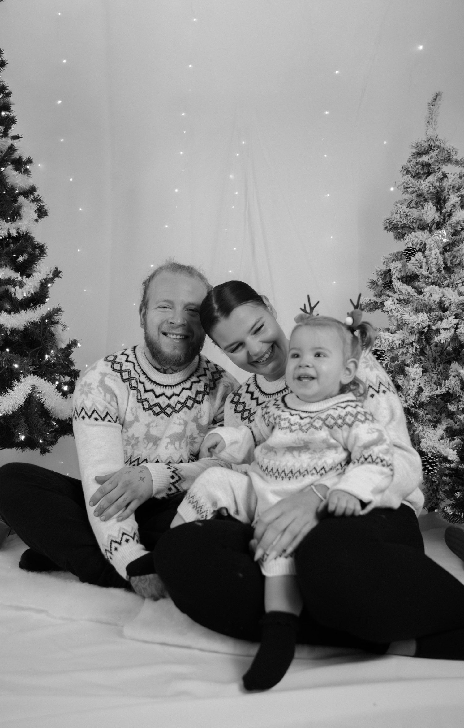 Portrait famille Essonne 91, parents et bébé en pulls de Noël assortis souriants devant sapins enneigés, séance photo Noël noir et blanc
