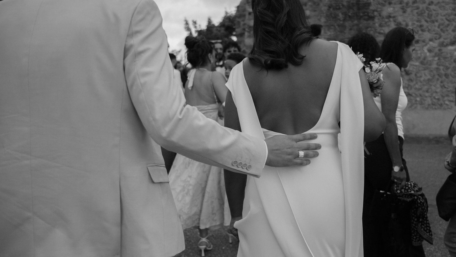 Photographe mariage Essonne 91, marié guidant tendrement sa femme dos nu en robe blanche fluide parmi les invités, noir et blanc élégant