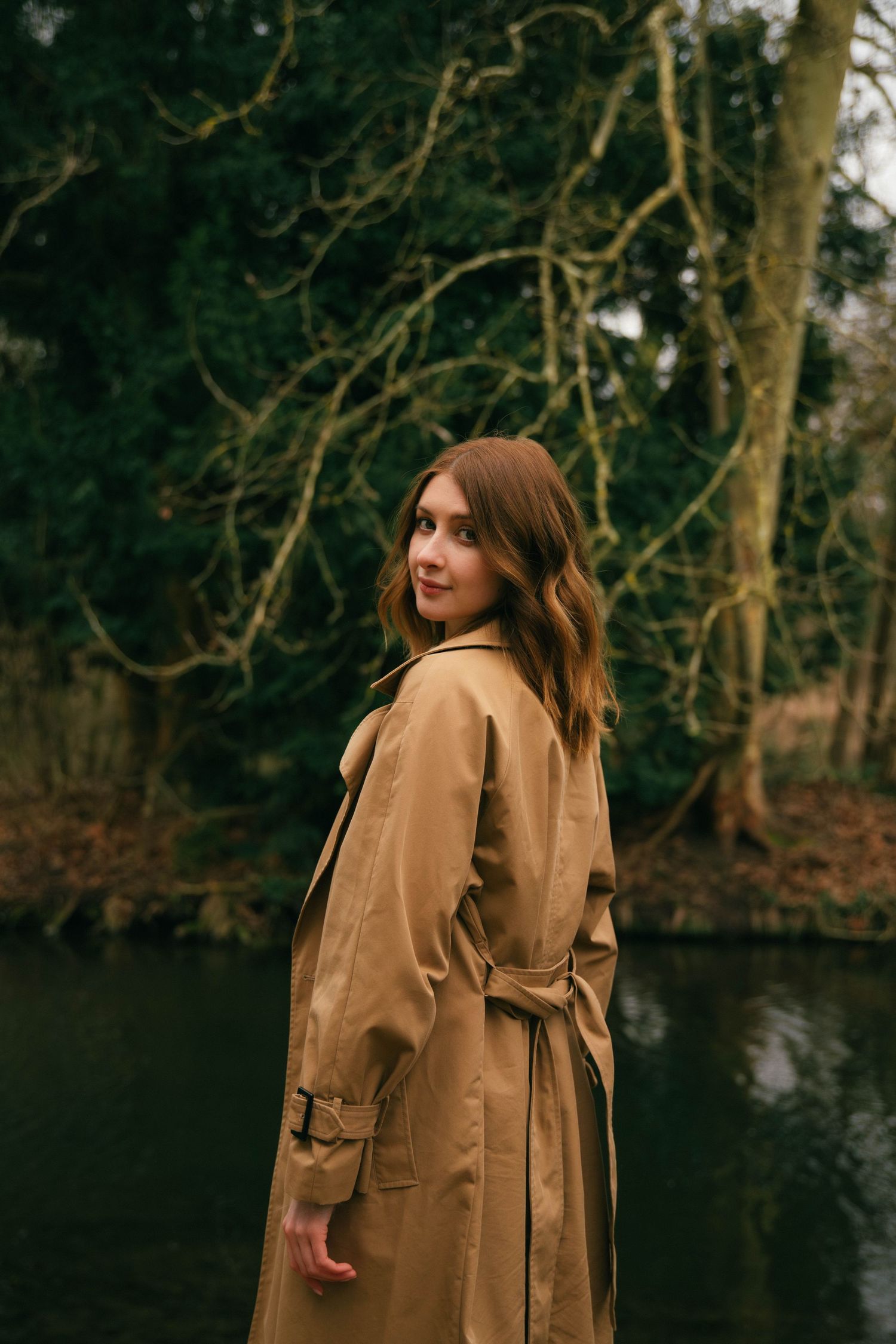 Portrait féminin Essonne 91, jeune femme en trench camel se retournant au bord d'un lac dans un parc boisé, séance photo lifestyle douce