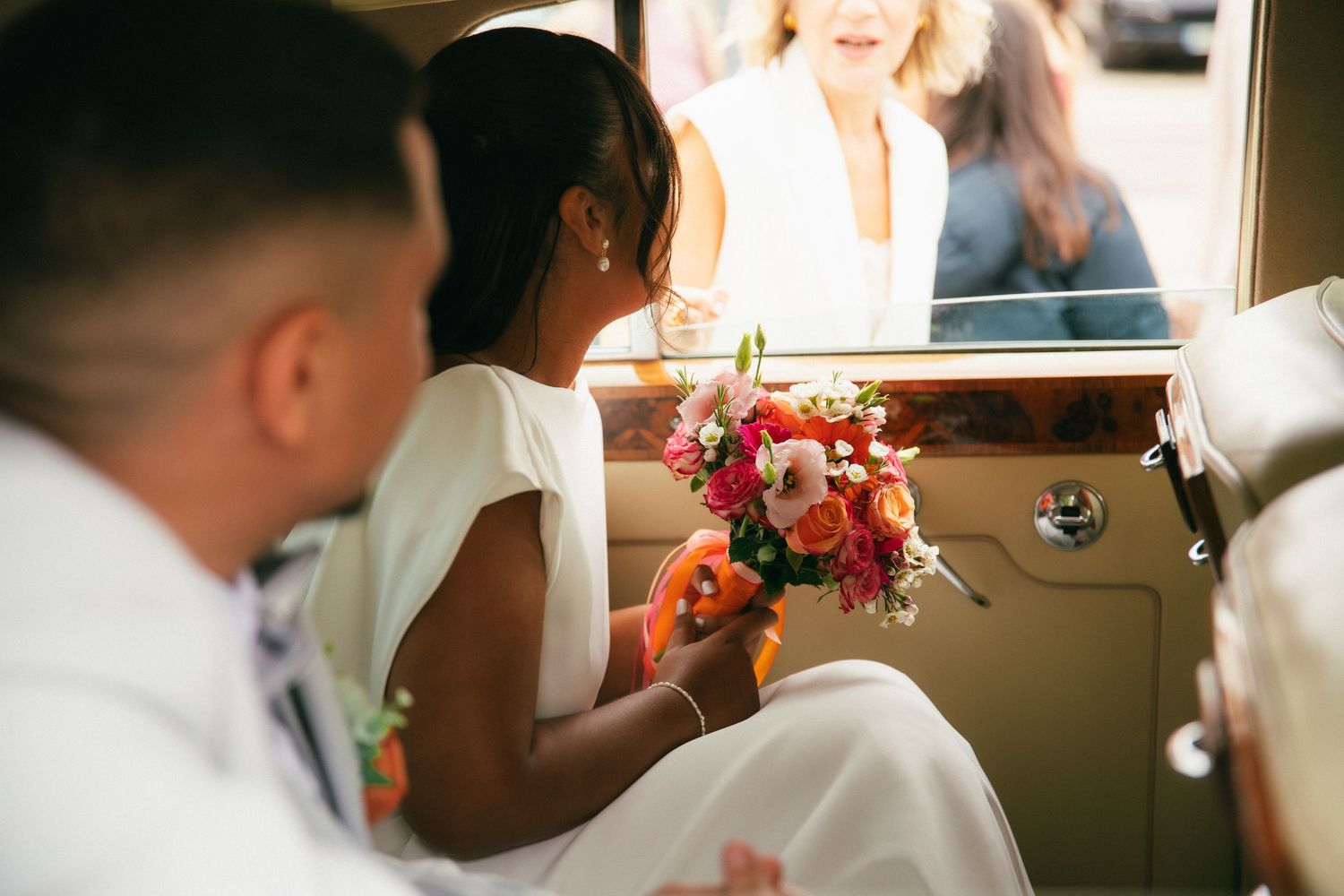 Une mariée élégamment vêtue tient son bouquet de fleurs aux teintes roses, orange et blanches tout en regardant par la fenêtre d'une voiture