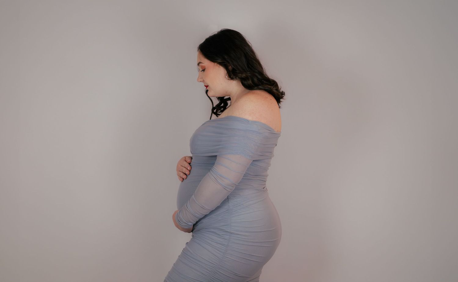 Portrait de profil d'une femme enceinte en robe bleue moulante sur fond gris clair. Séance grossesse en studio Essonne.