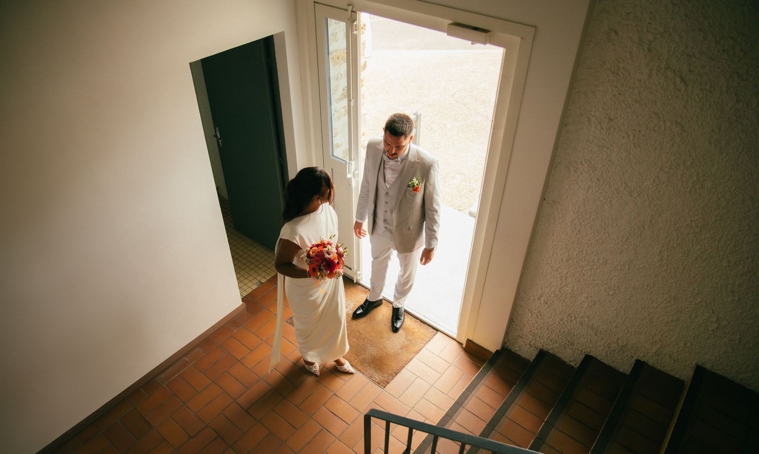Vue en plongée des mariés entrant dans un bâtiment, bouquet de roses rouges en main. Reportage mariage en Essonne. Photographe mariage 91.