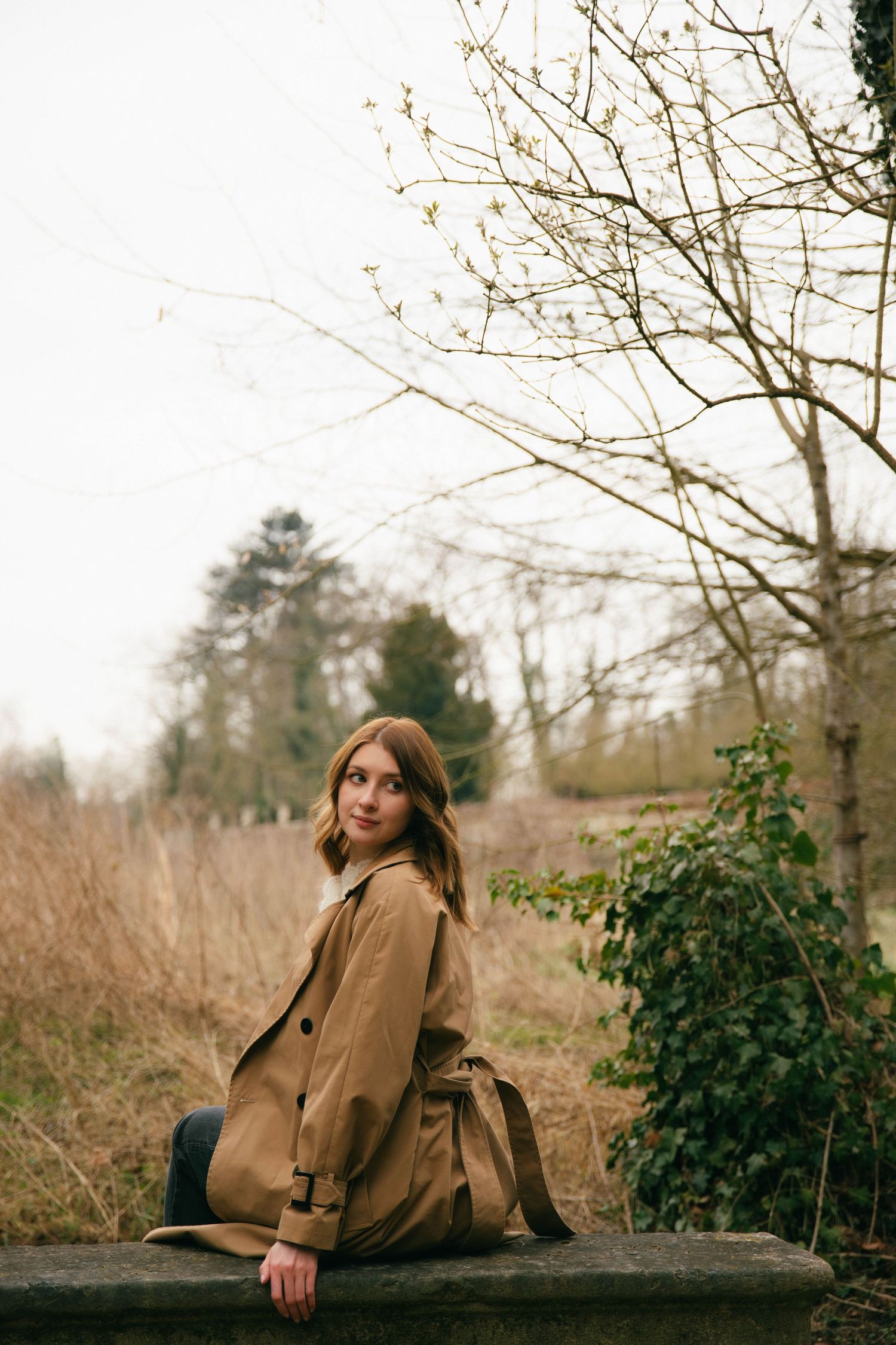 Portrait d'une femme assise sur un muret en trench camel, regard naturel dans un parc en hiver. Séance portrait en Essonne. Photographe 91.