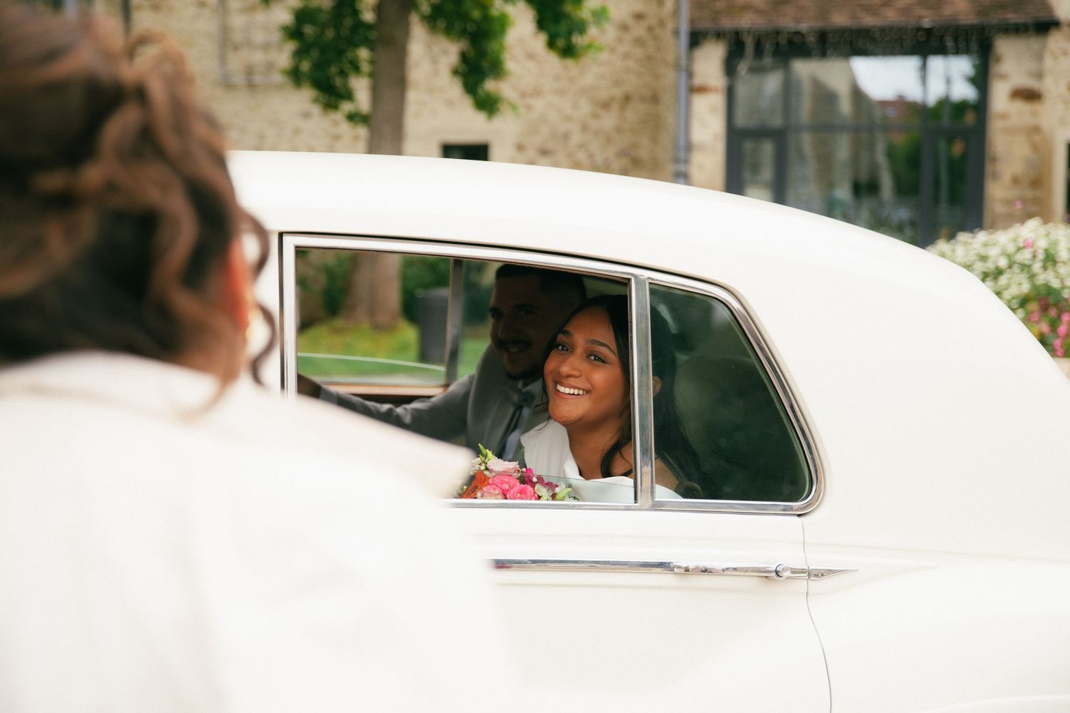 Mariée souriante à bord d'une voiture de collection blanche, bouquet coloré en main. Reportage mariage en Essonne. Photographe mariage 91.