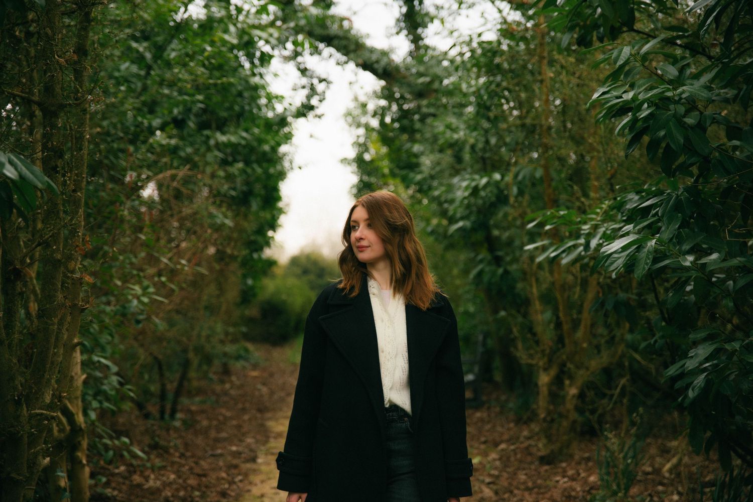 Photographe Essonne 91, portrait jeune femme en manteau noir marchant dans une allée verdoyante, photo lifestyle naturelle et poétique