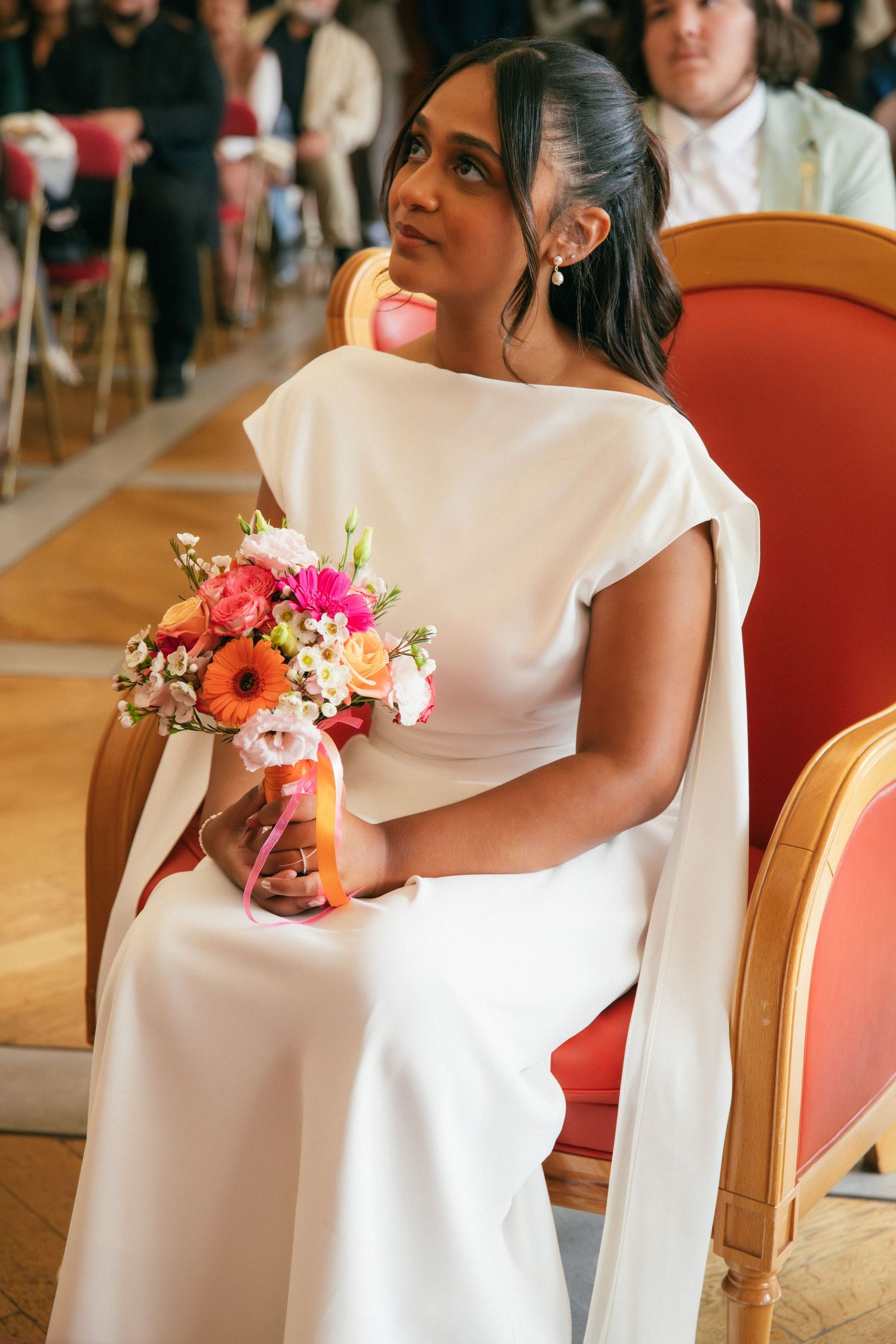 Photographe mariage Essonne 91, mariée élégante assise en robe blanche fluide tenant un bouquet de fleurs colorées lors de la cérémonie