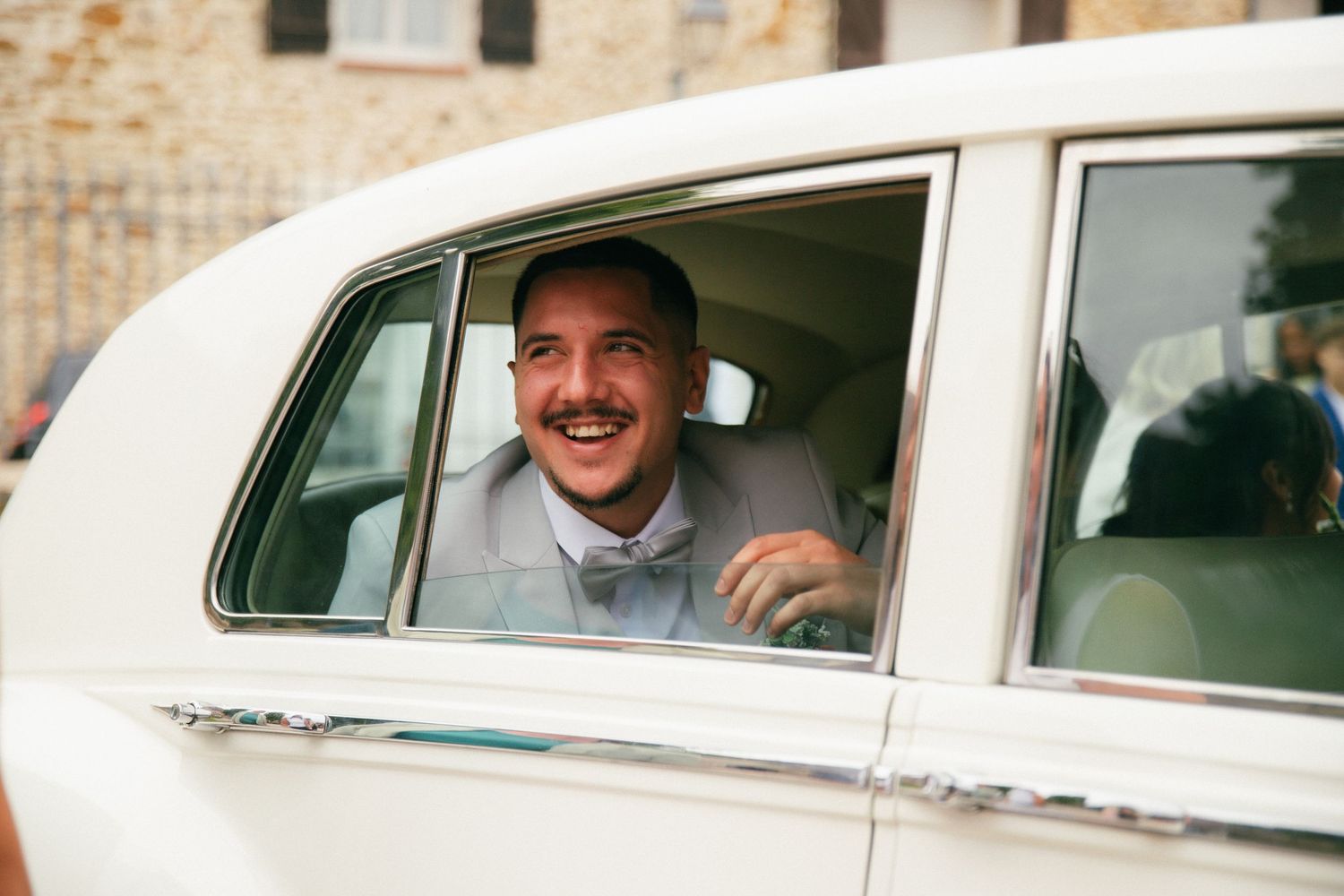 Photographe mariage Essonne 91, marié souriant et complice à la fenêtre d'une voiture de collection blanche, portrait spontané