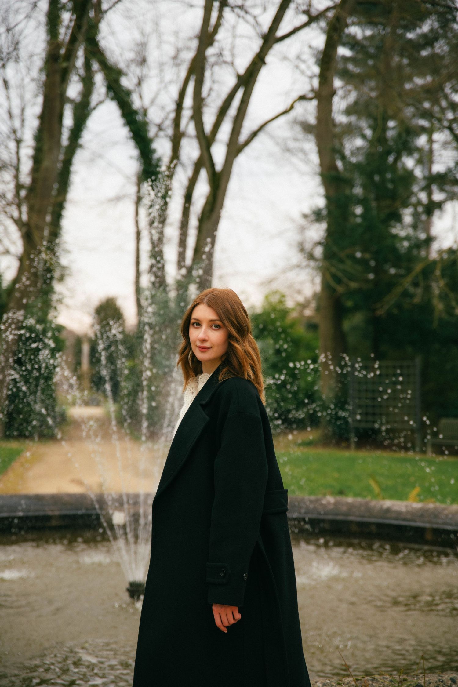 Séance photo lifestyle Essonne 91, jeune femme en long manteau noir devant une fontaine dans un parc, portrait féminin élégant et naturel