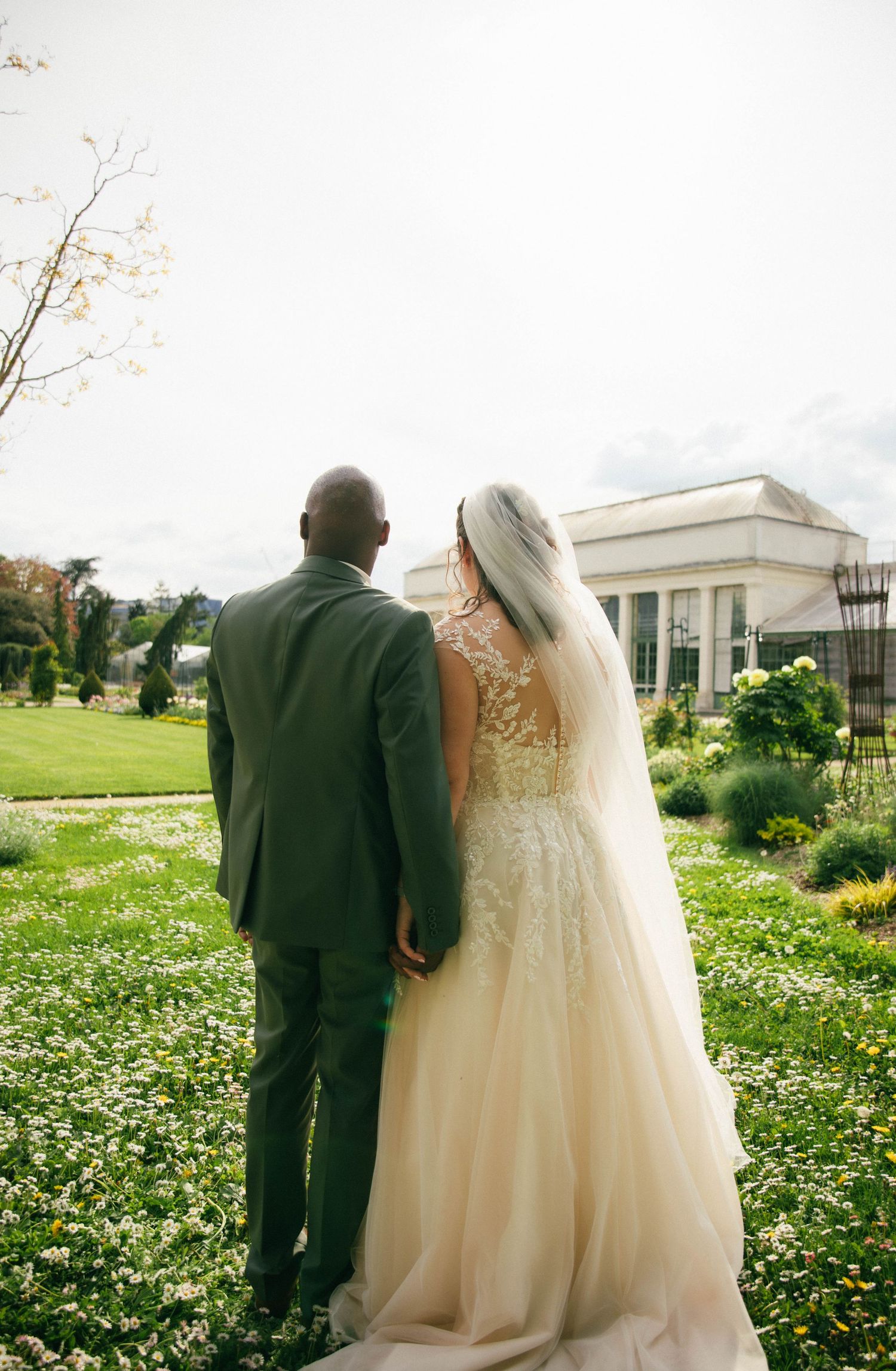 Photographe mariage Essonne 91, mariés de dos contemplant un domaine fleuri, robe de mariée en dentelle et voile, reportage mariage élégant
