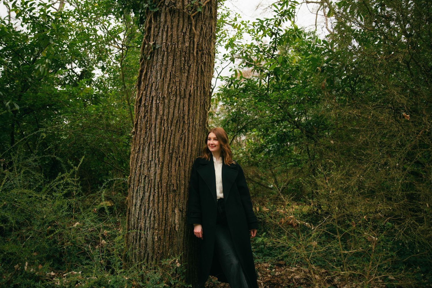 Portrait d'une femme adossée à un arbre en forêt, manteau noir et végétation luxuriante. Séance portrait en extérieur Essonne. Photographe
