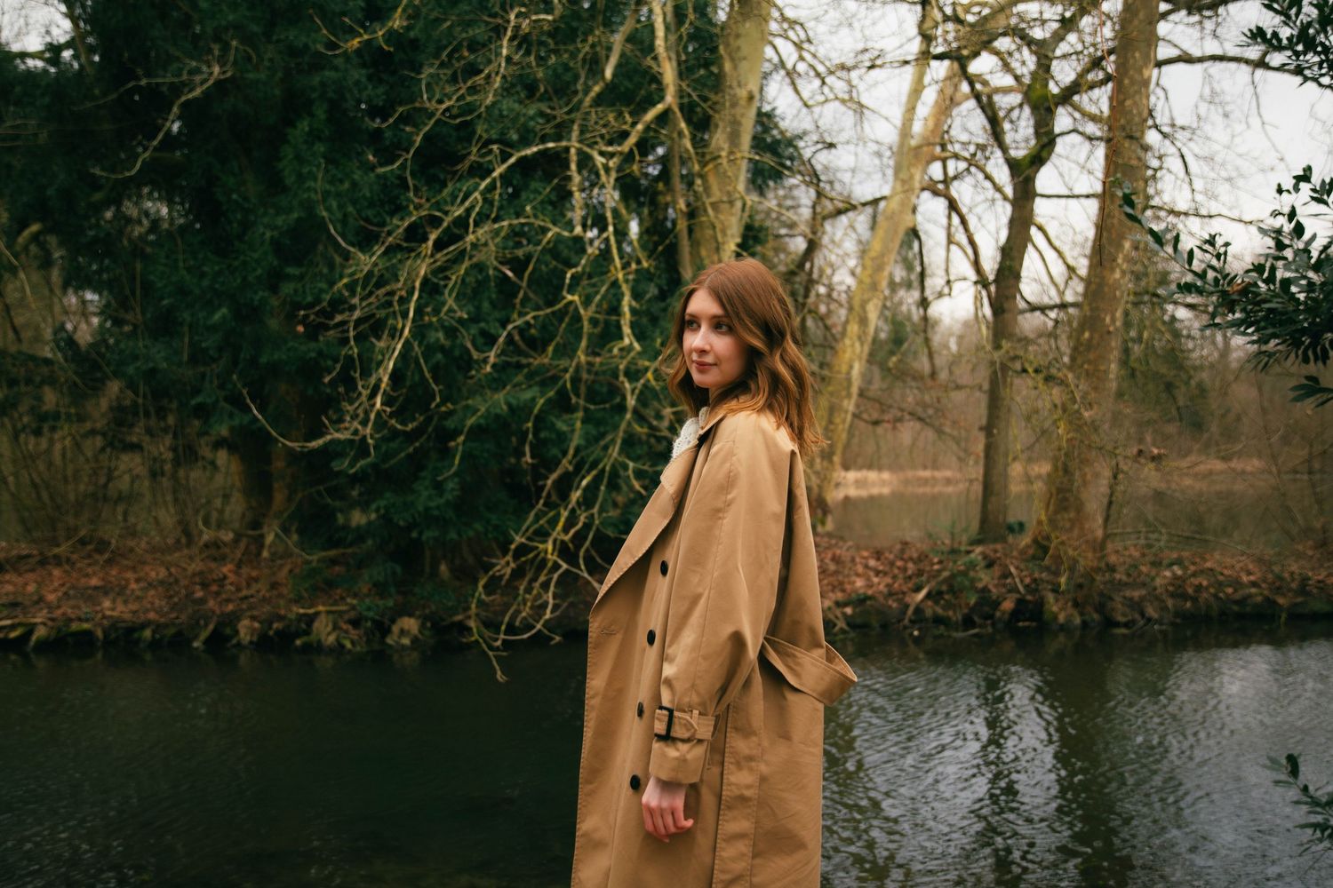 Photographe Essonne 91, portrait jeune femme en trench camel se retournant au bord d'un lac en forêt hivernale, séance photo lifestyle