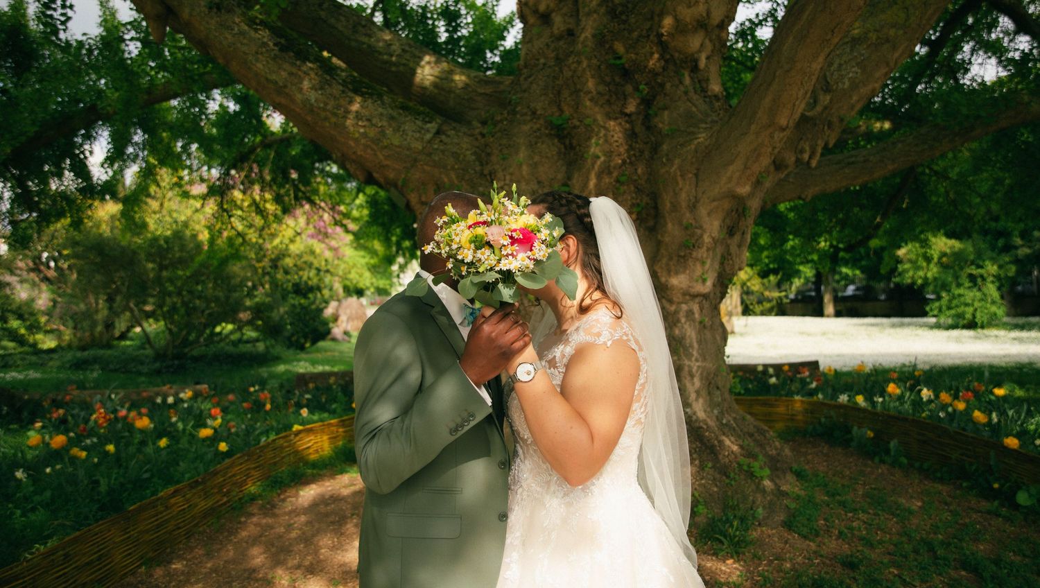 Photographe mariage Essonne 91, mariés s'embrassant sous un grand arbre fleuri, bouquet de mariée coloré, reportage mariage romantique et na