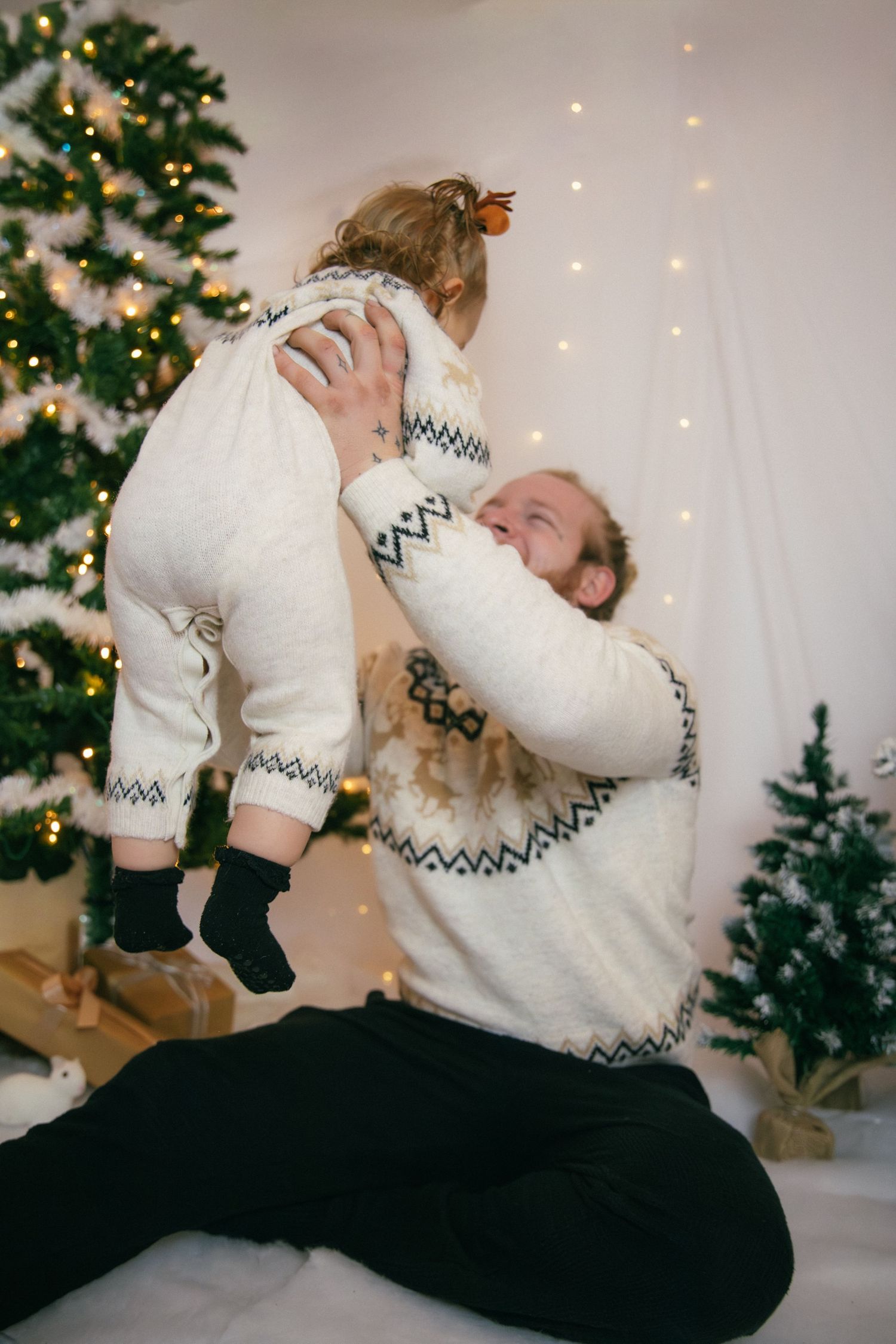 Séance photo Noël Essonne 91, papa soulevant son bébé en l'air avec tendresse devant un sapin illuminé, portrait famille joyeux et complice