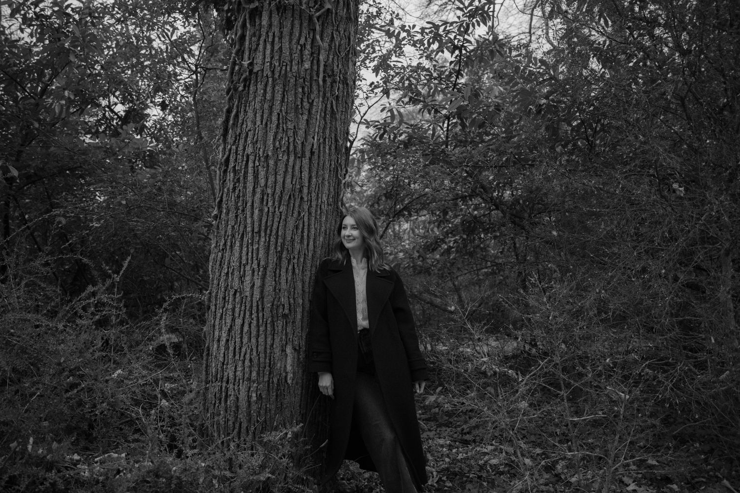 Portrait en noir et blanc d'une femme adossée à un grand arbre en forêt, atmosphère sombre et élégante. Séance photo Essonne, 91.