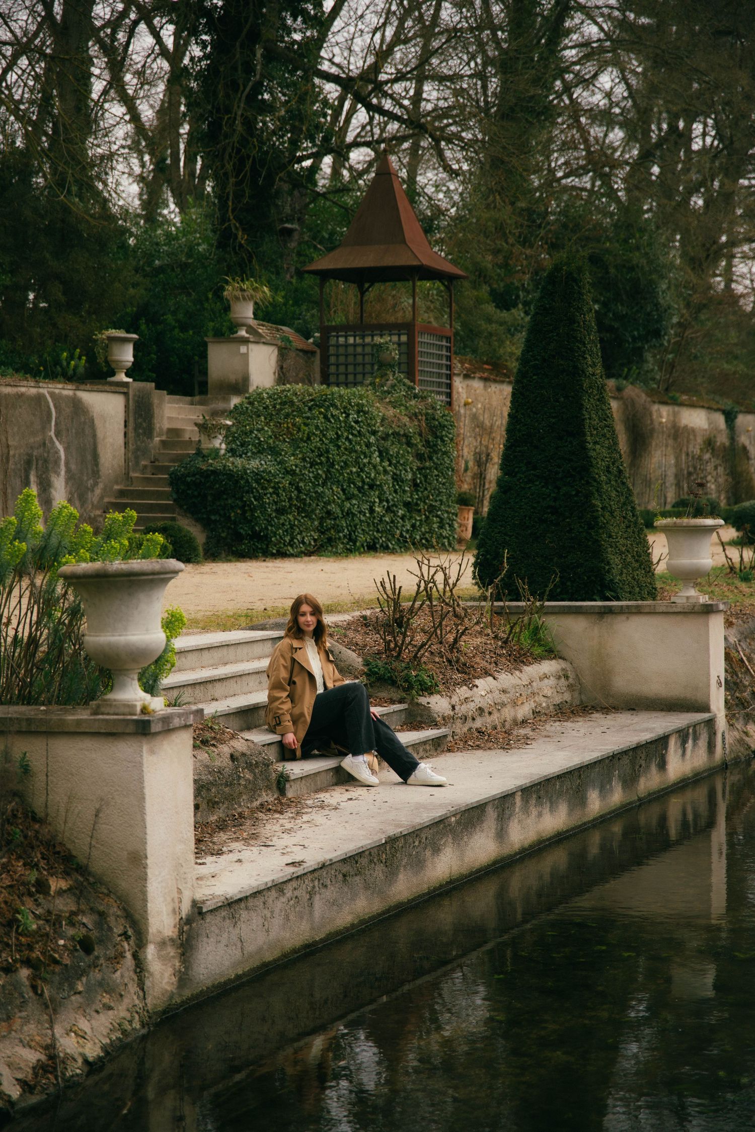 Photographe Essonne 91, portrait jeune femme en trench camel assise au bord d'un bassin dans un parc à la française, séance photo lifestyle