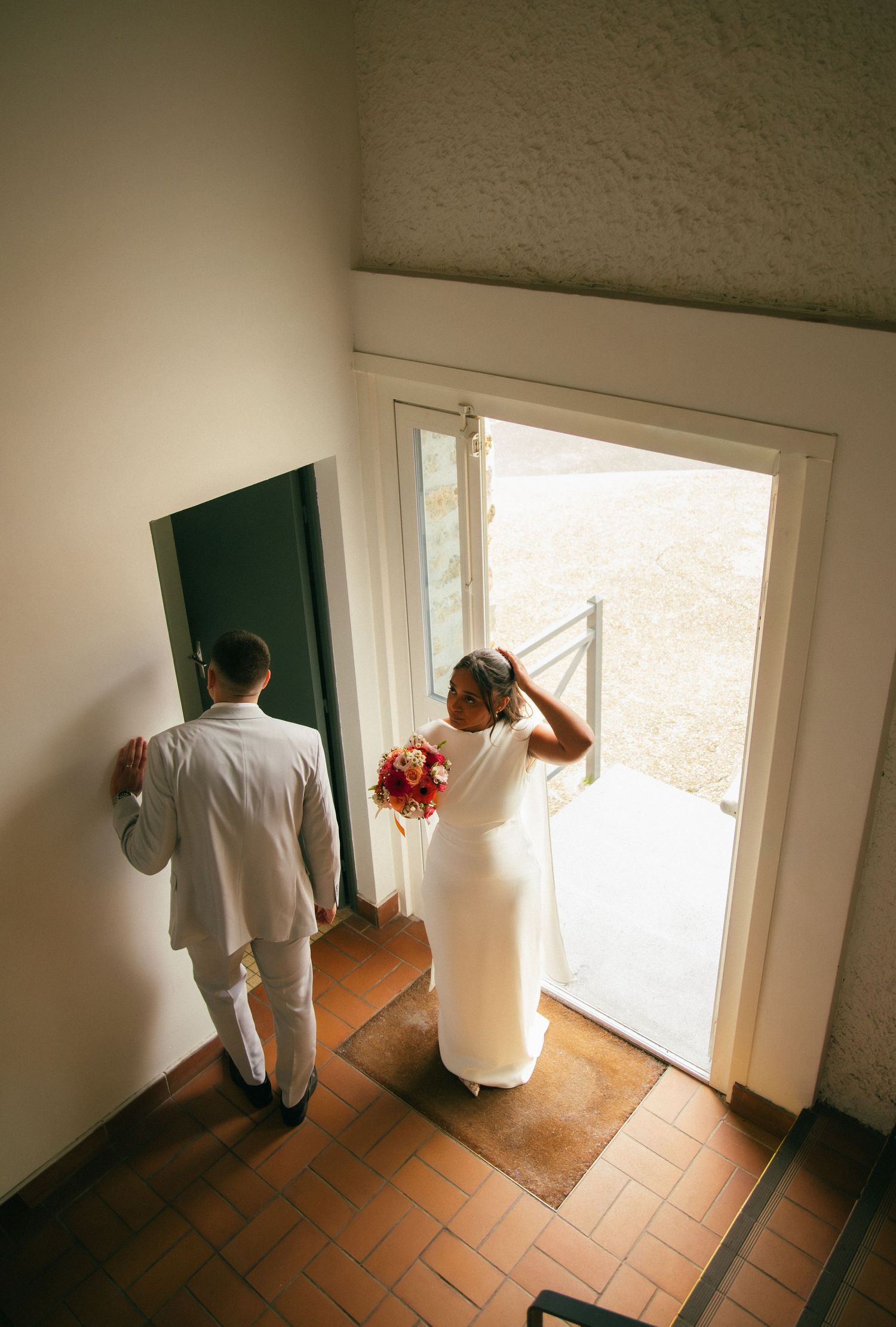 Reportage mariage Essonne 91, mariés vus en plongée s'apprêtant à sortir vers la lumière, robe blanche et bouquet coloré, instant émouvant