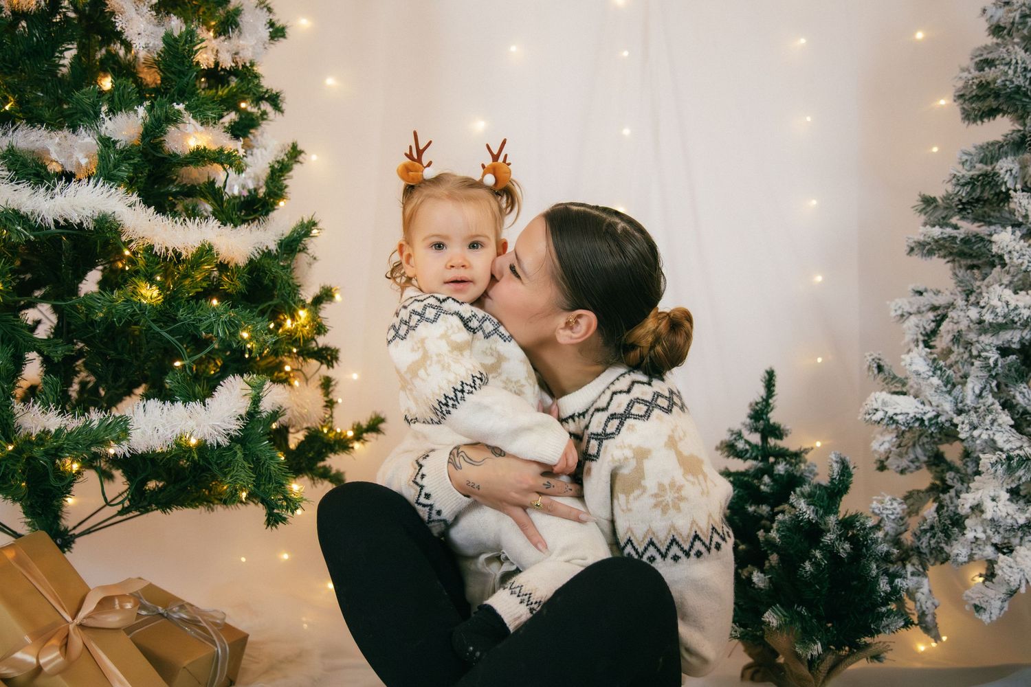 Séance photo Noël Essonne 91, maman et bébé en pulls assortis riant ensemble devant un sapin enneigé, portrait famille chaleureux et festif