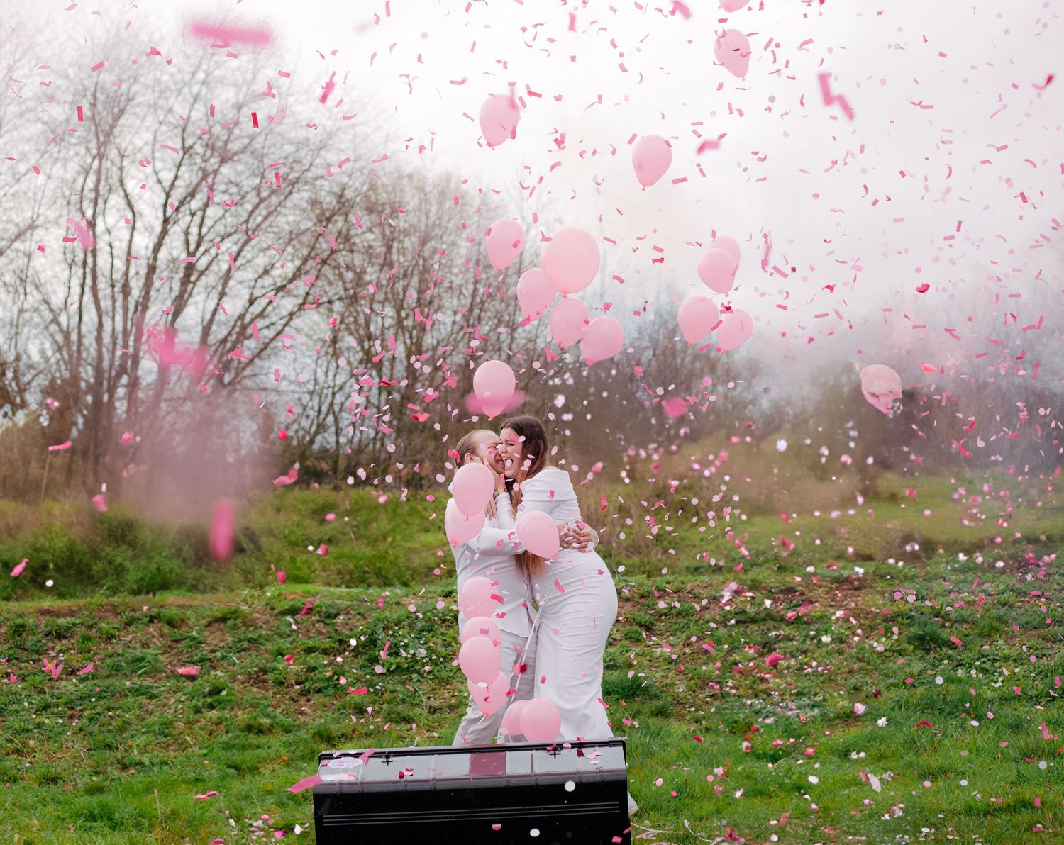 Couple s'embrassant sous une explosion de confettis et ballons roses lors d'un gender reveal en plein air. Photographe événement Essonne 91.