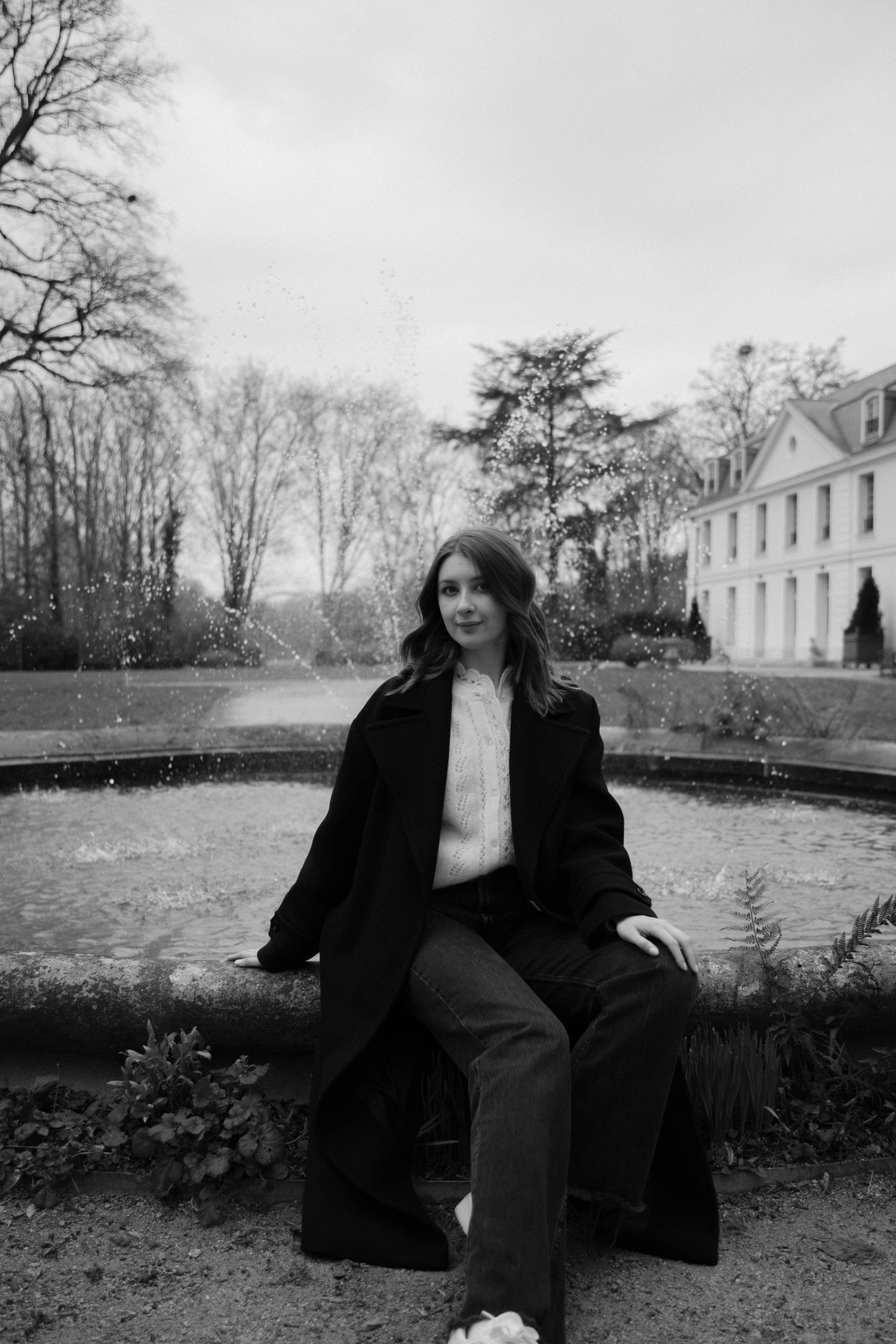 Portrait féminin Essonne 91, jeune femme en manteau noir assise devant une fontaine et demeure bourgeoise, noir et blanc, séance photo life