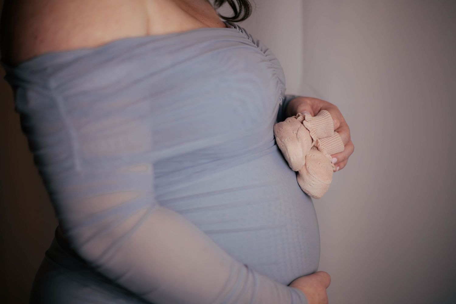 Gros plan sur le ventre d'une femme enceinte en robe bleue tenant de petits chaussons roses. Séance grossesse Essonne. Photographe maternité