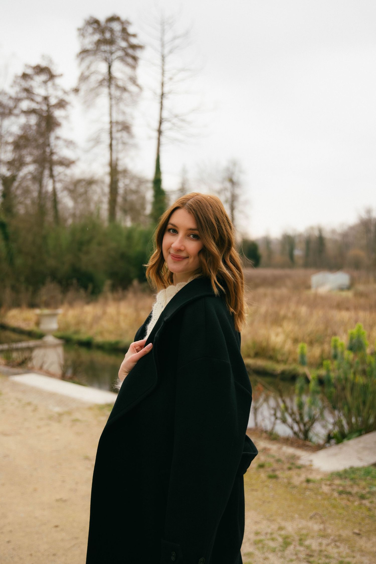 Portrait d'une femme en manteau noir au bord d'un étang en hiver, regard naturel et atmosphère douce.Séance portrait en Essonne. Photographe