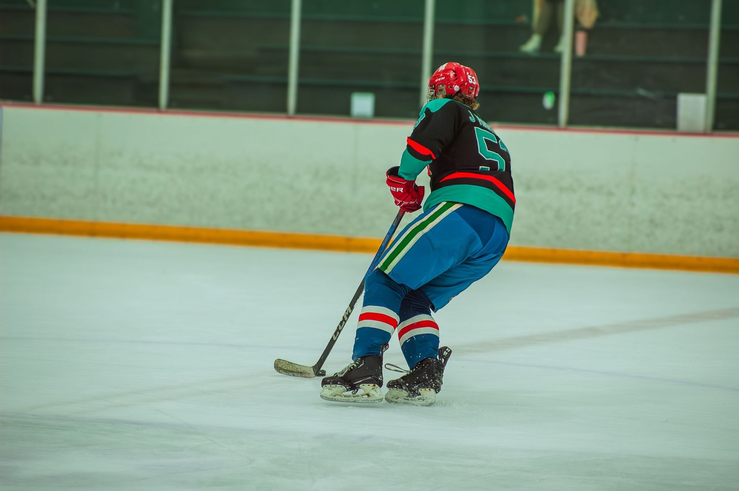 A hockey player in a teal and red uniform skates with a stick during an indoor ice hockey game.