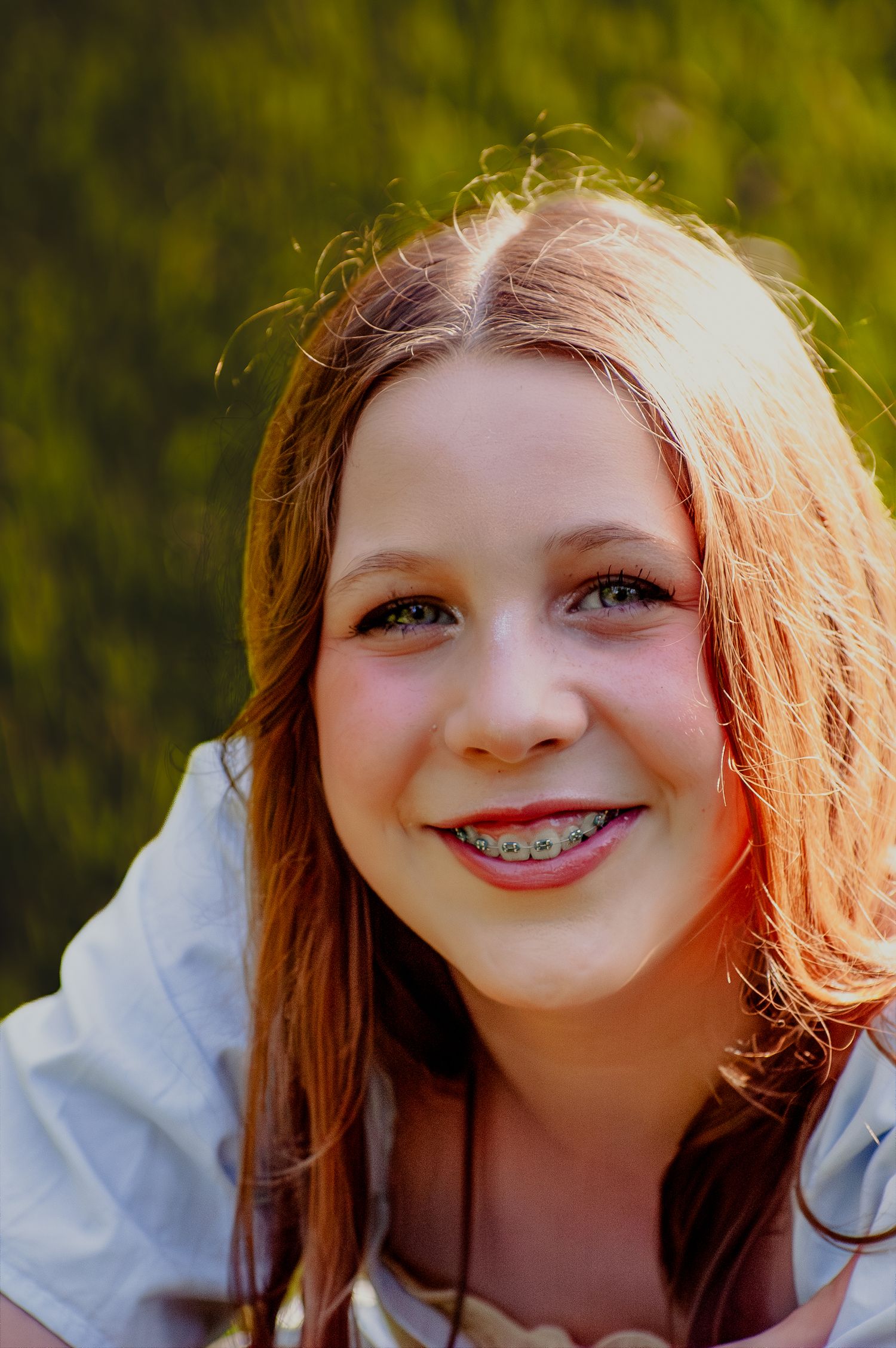 Smiling teenage girl with braces and long auburn hair posing outdoors in warm sunlight.