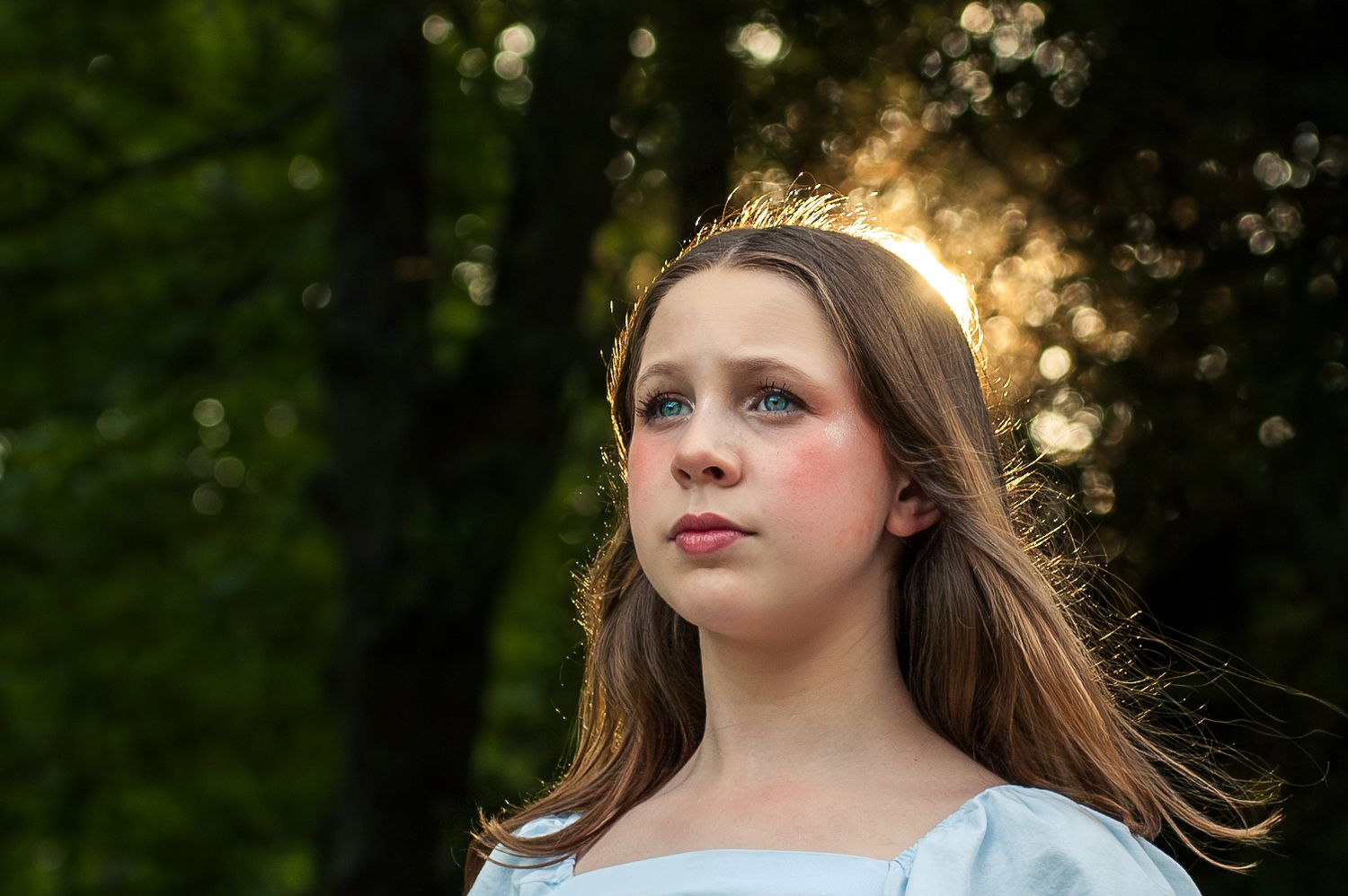 Young girl wearing a gold crown headband in a blue dress with sunlight glowing behind her.