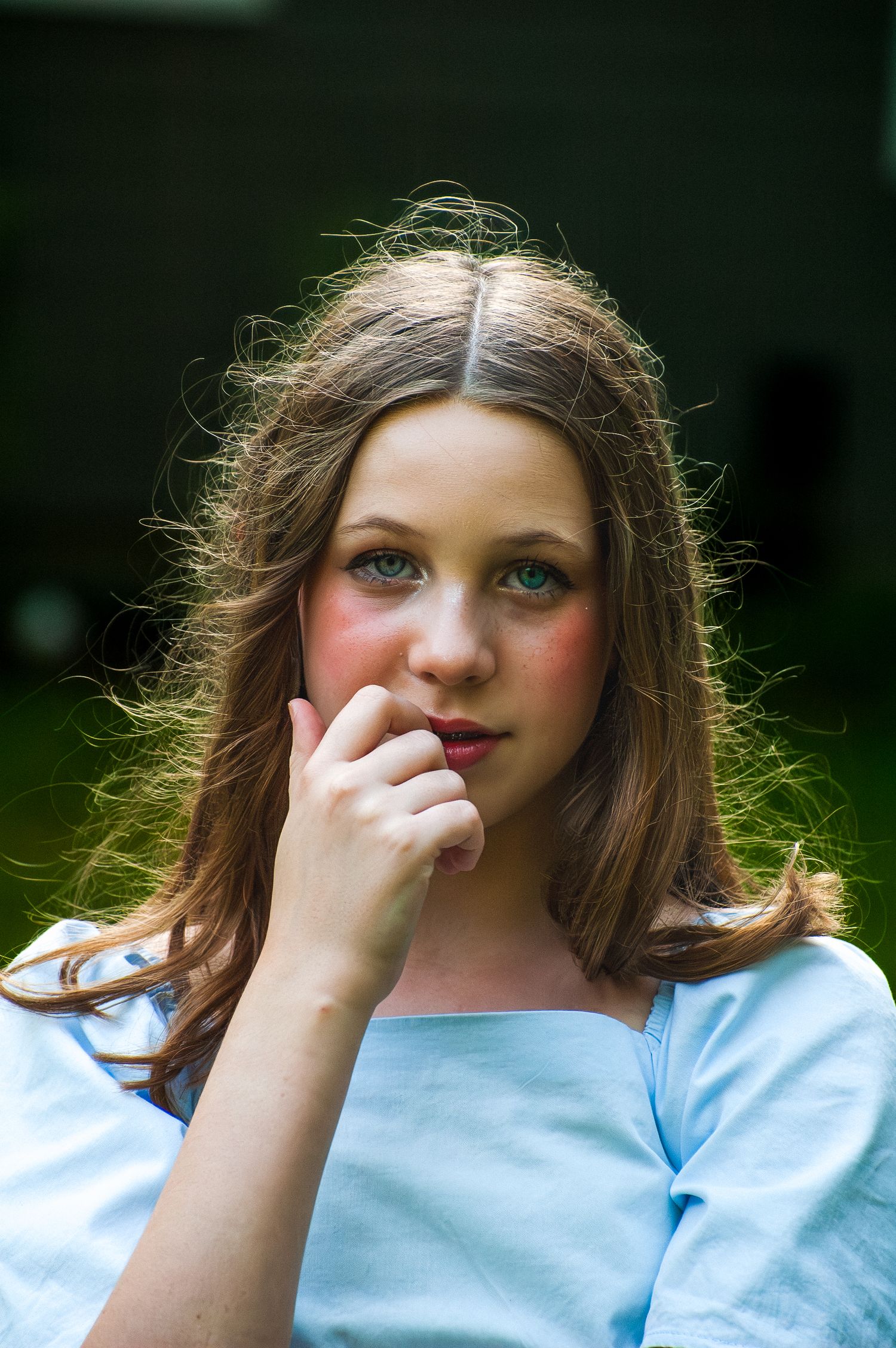 A young woman with long brown hair and blue eyes in a light blue top poses outdoors thoughtfully.
