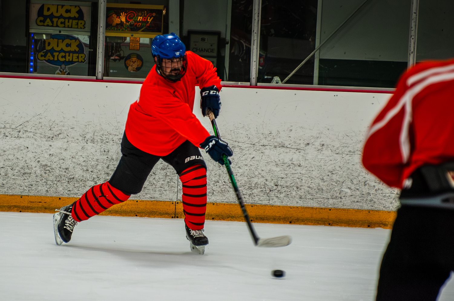 Hockey player in red jersey and blue helmet shooting puck on indoor ice rink.