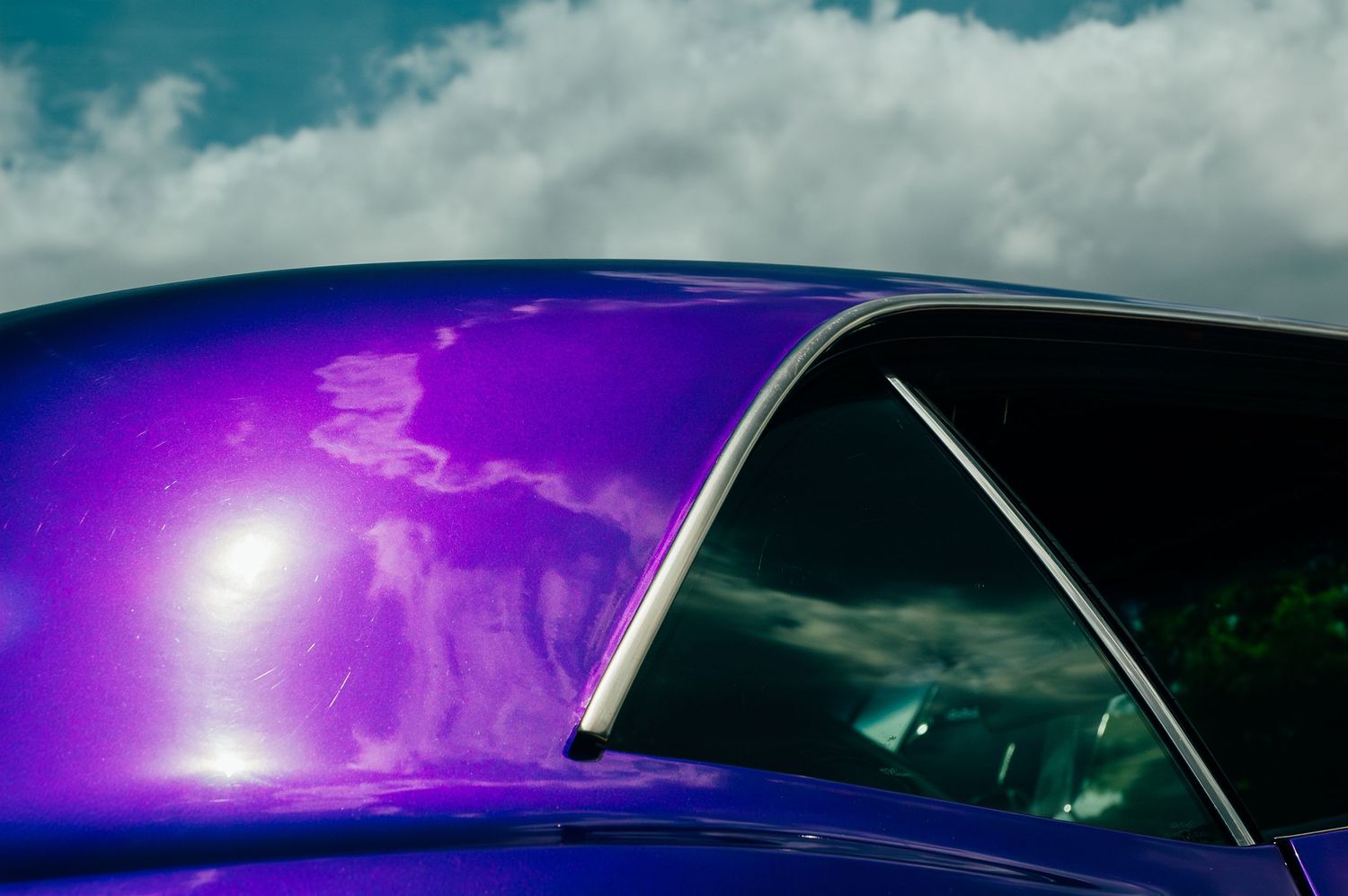 Close-up of a shiny purple classic car roof reflecting dramatic clouds against a blue sky.