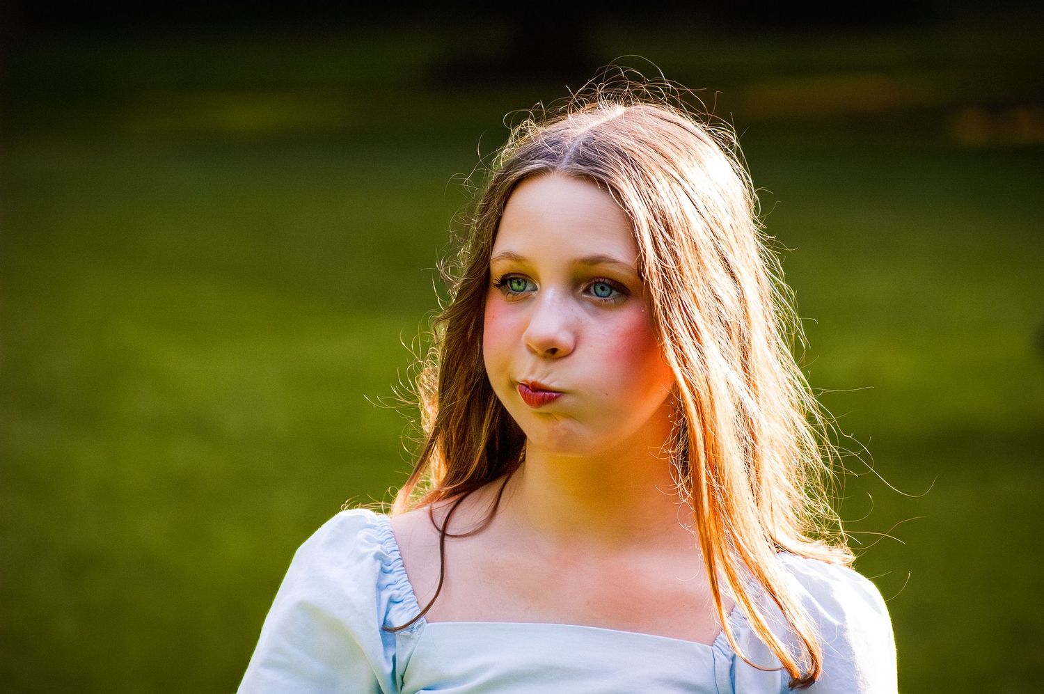 Young girl with long brown hair wearing a light blue dress posing outdoors on green grass.