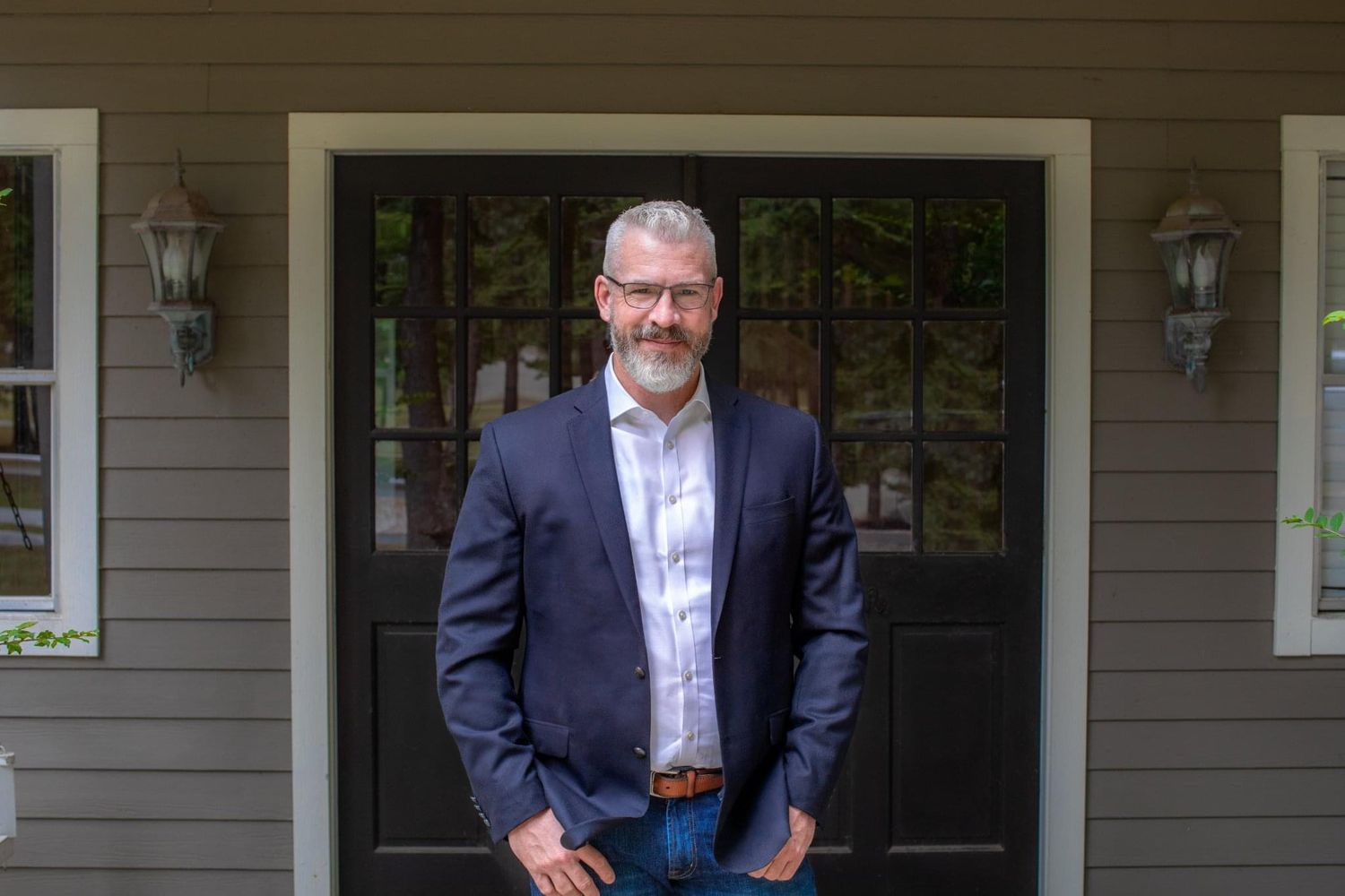 A professional man in a navy blazer and white shirt stands confidently in front of a dark door of a home.