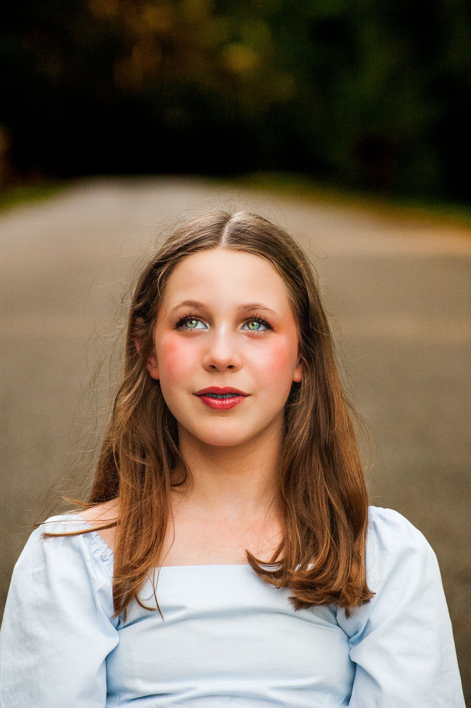 Young teenage girl with green eyes and brown hair in a light blue top looking upward outdoors.
