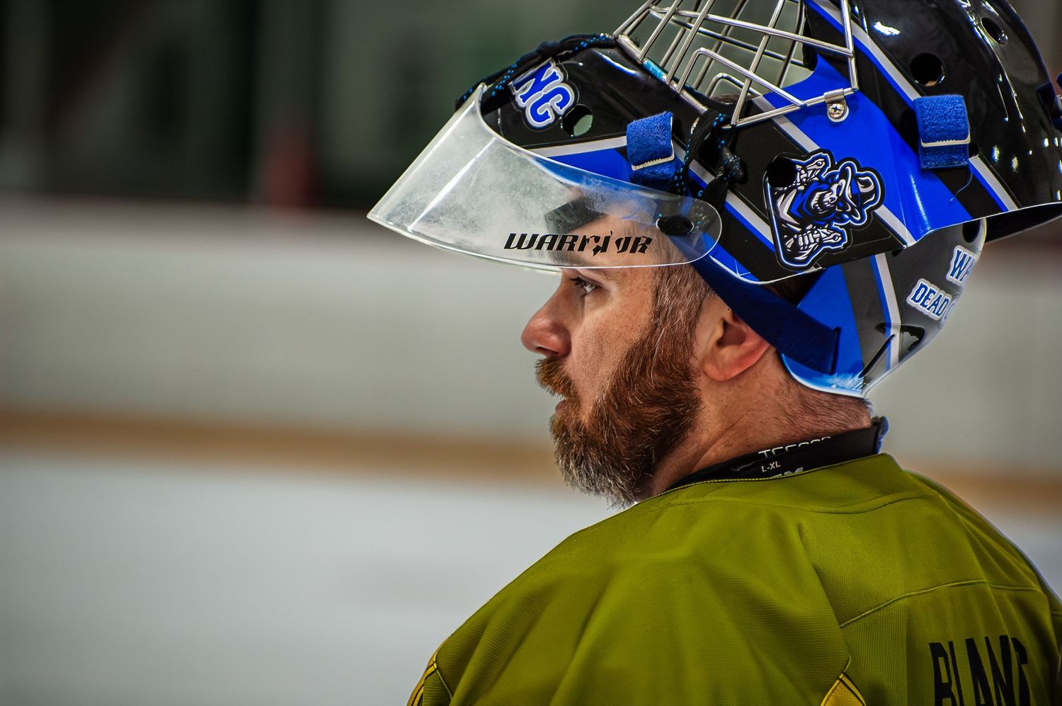 Bearded goalie wearing blue and black mask with olive green jersey during hockey game.